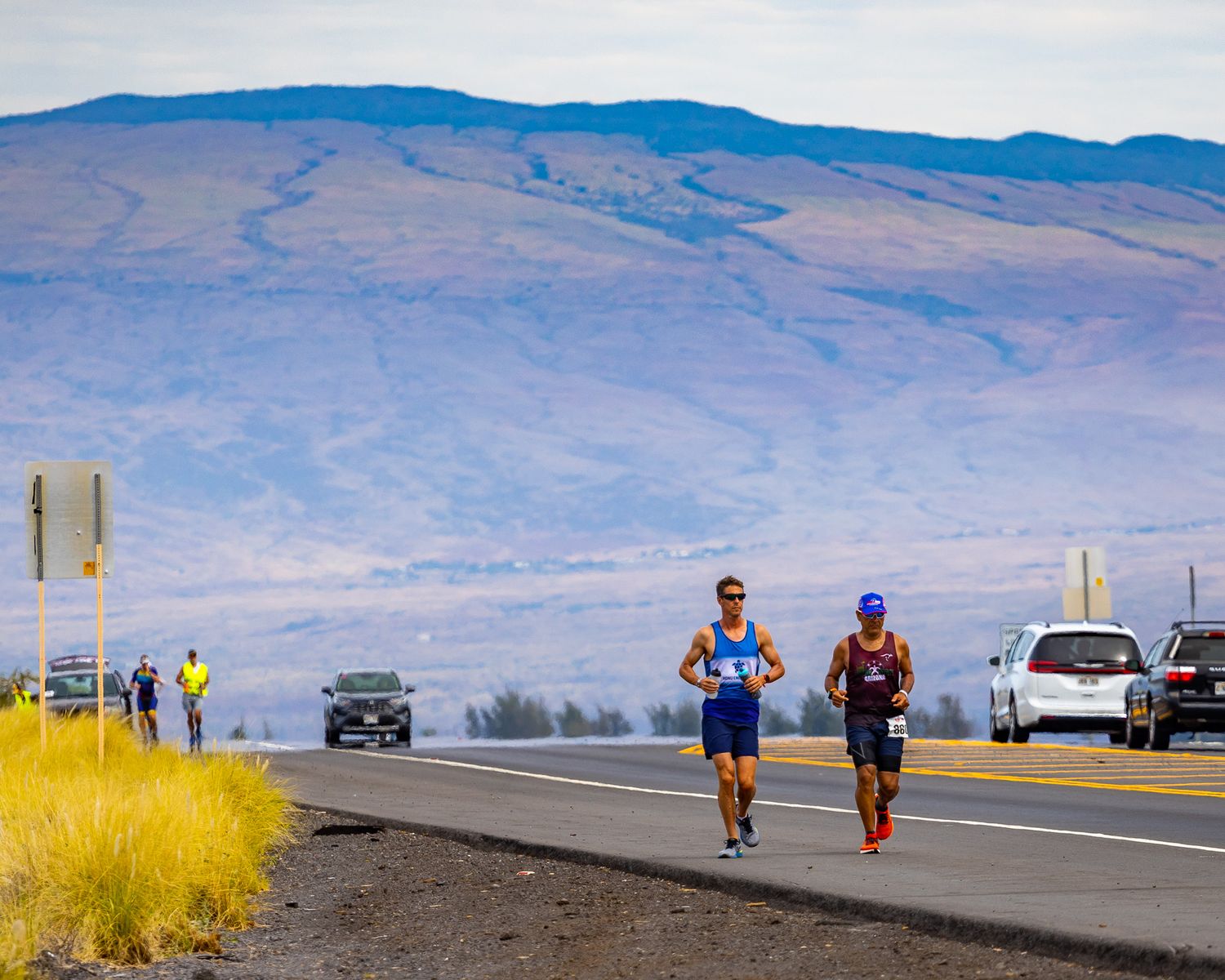 Two runners on an empty road with mountains and desert landscape in the background.