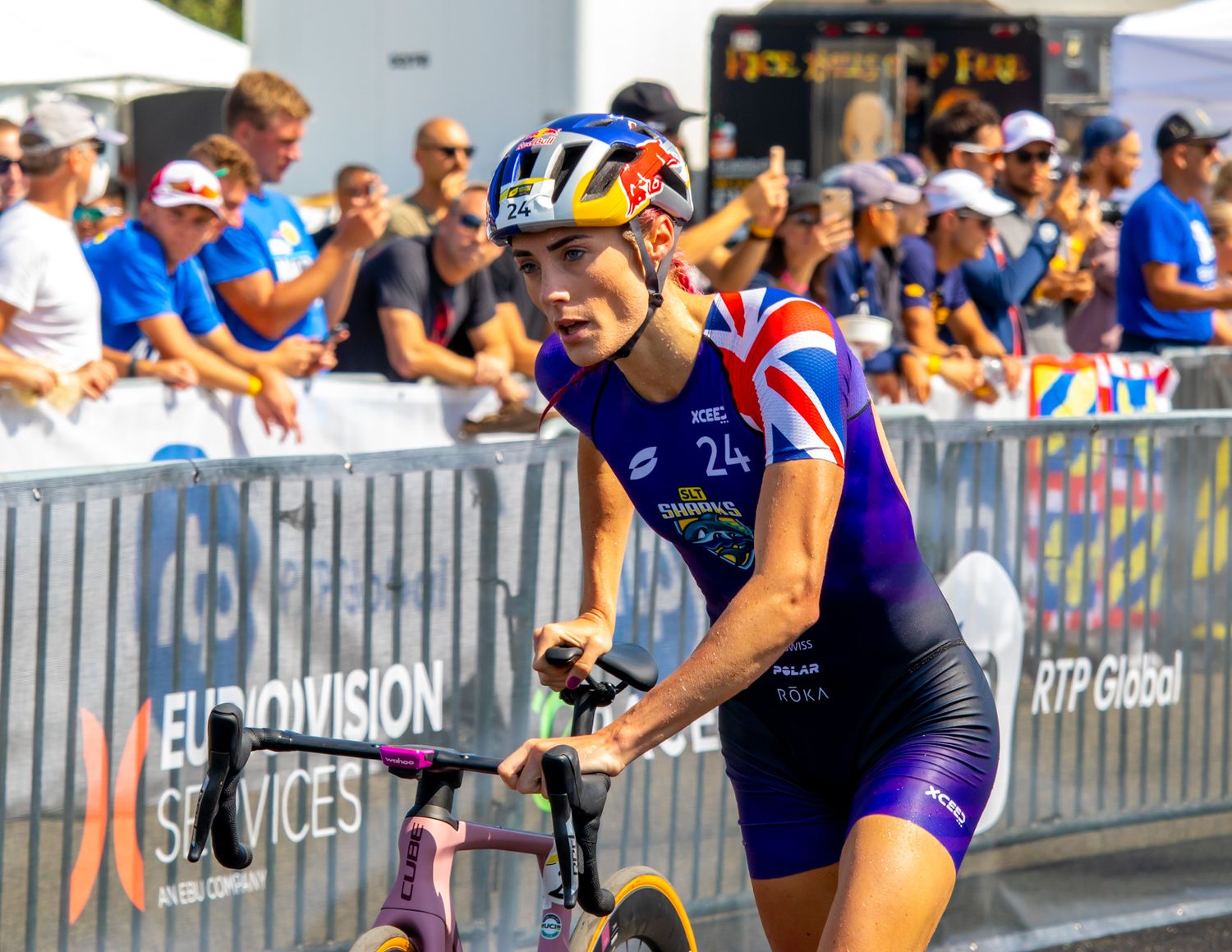 Cyclist in blue and red uniform competing in a triathlon event with spectators behind barriers.