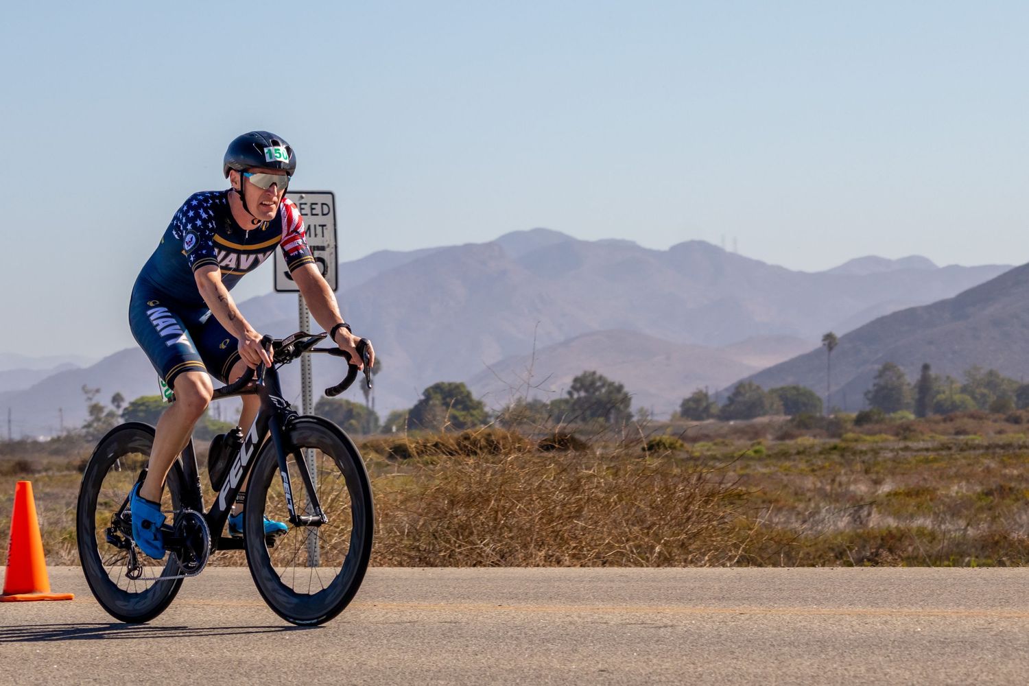 Cyclist in dark gear rides on mountain road with desert hills in background.