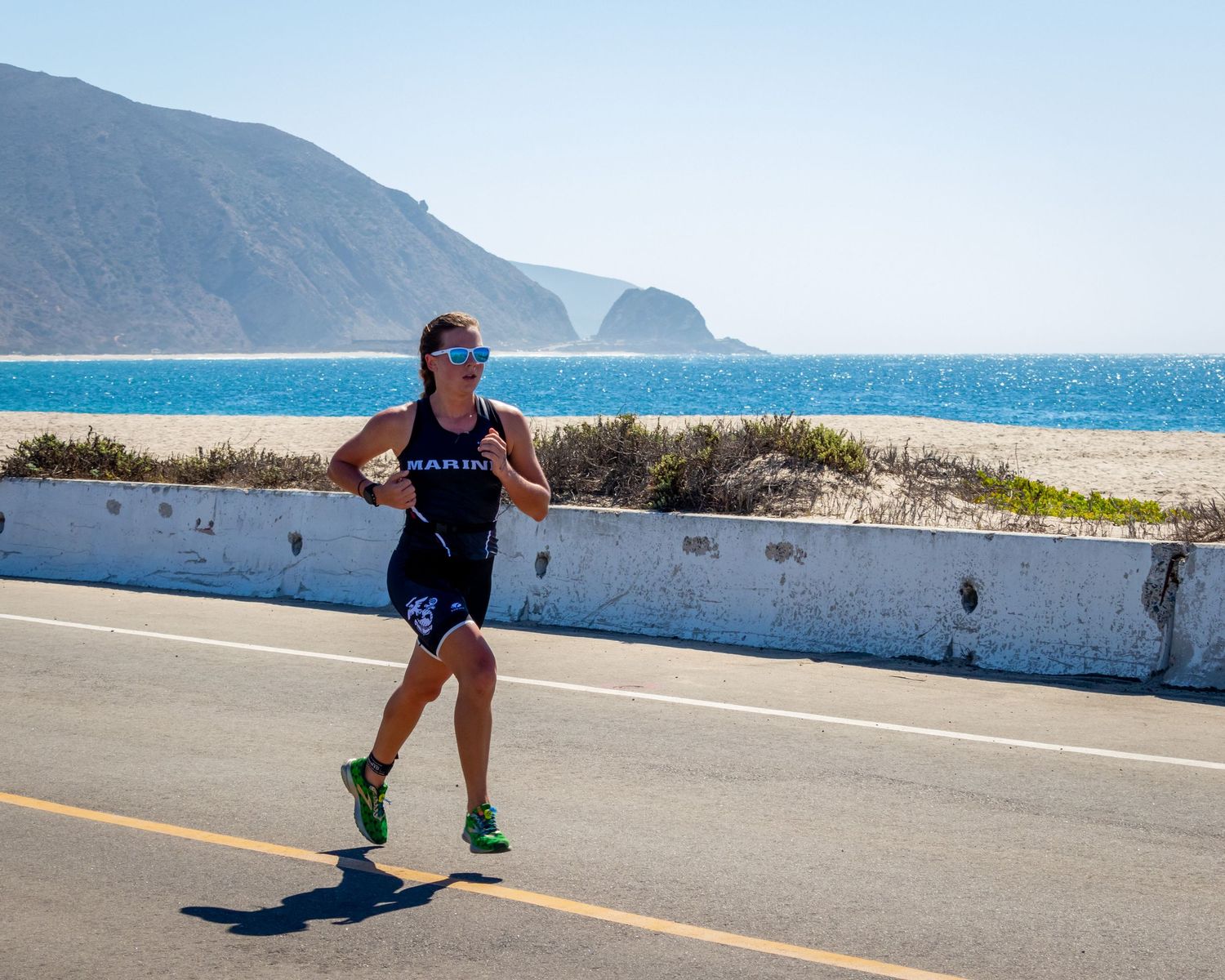 Triathlete running along coastal road with ocean and mountains in background.