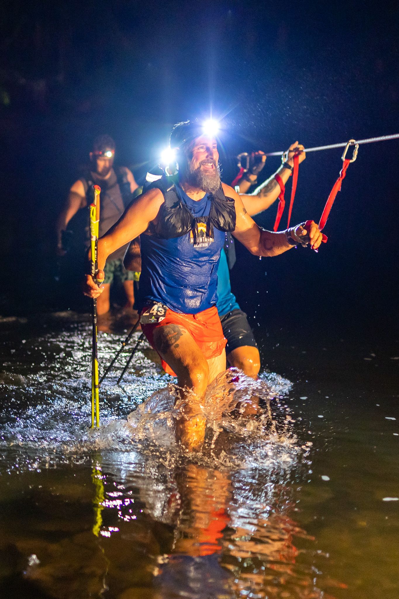 Runner splashing through water at night during an adventure race with headlamps illuminating the scene.