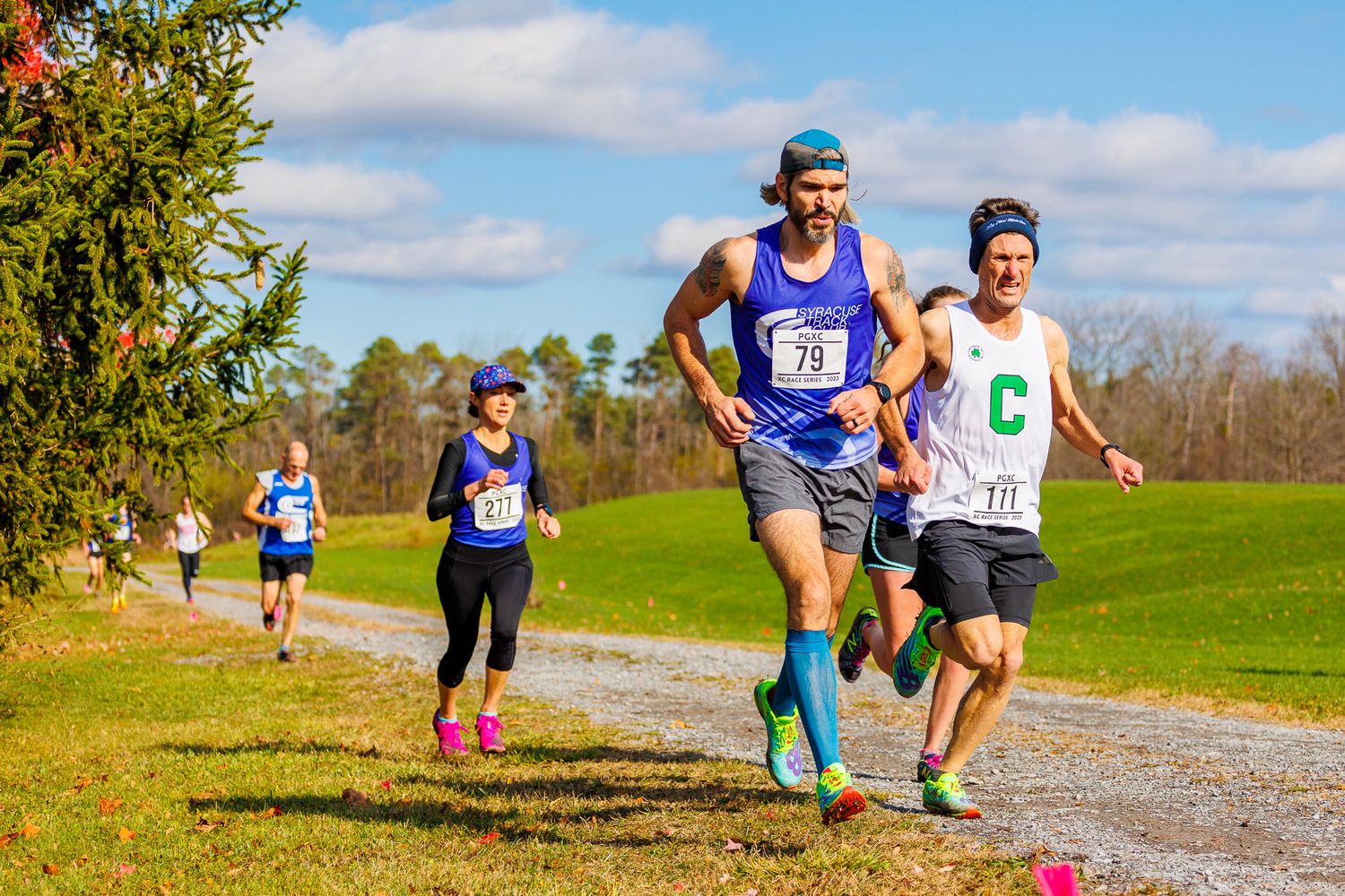 Runners racing along a dirt trail through grassy fields on a sunny day.