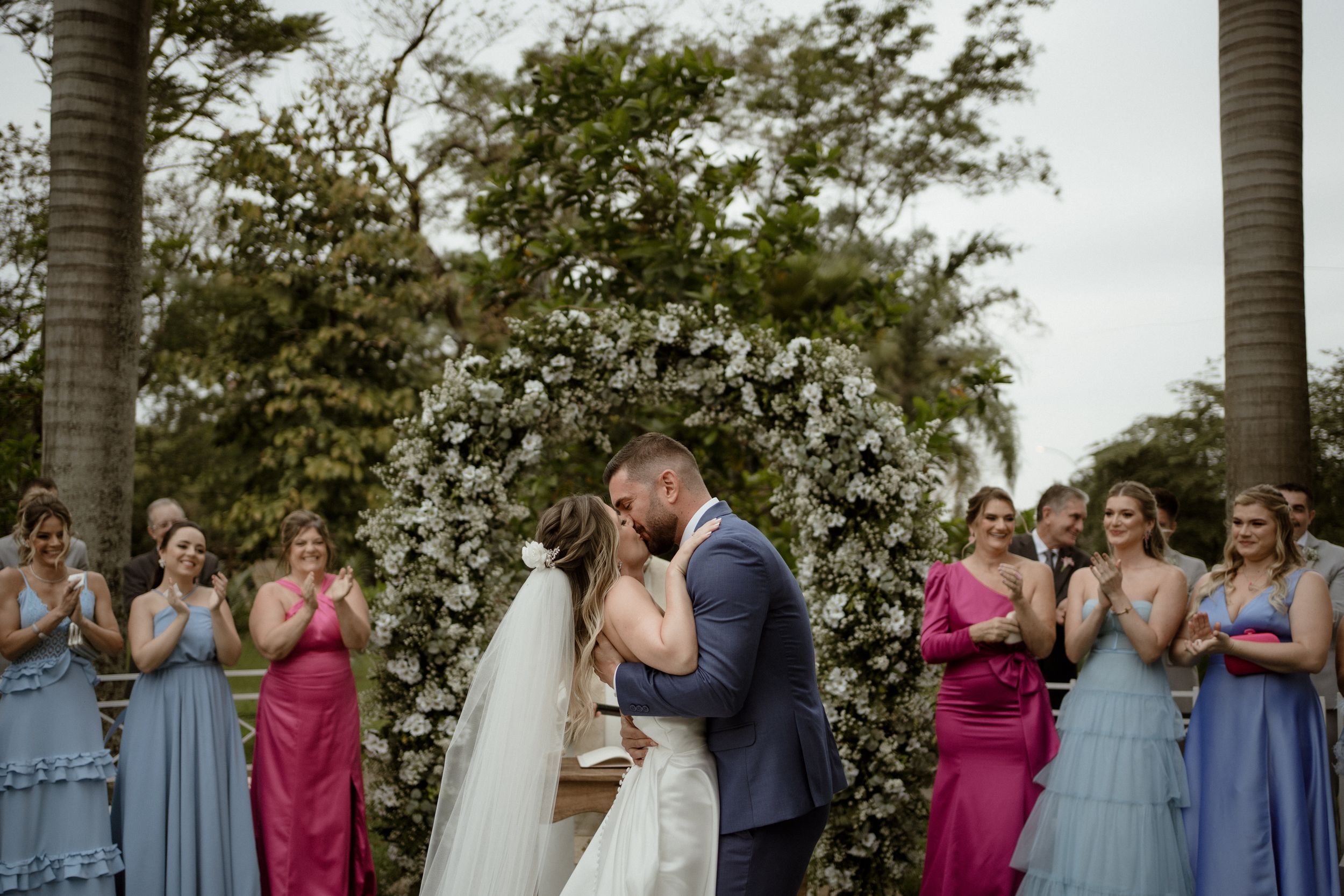 Wedding guests in colorful dresses watching the ceremony from white chairs.