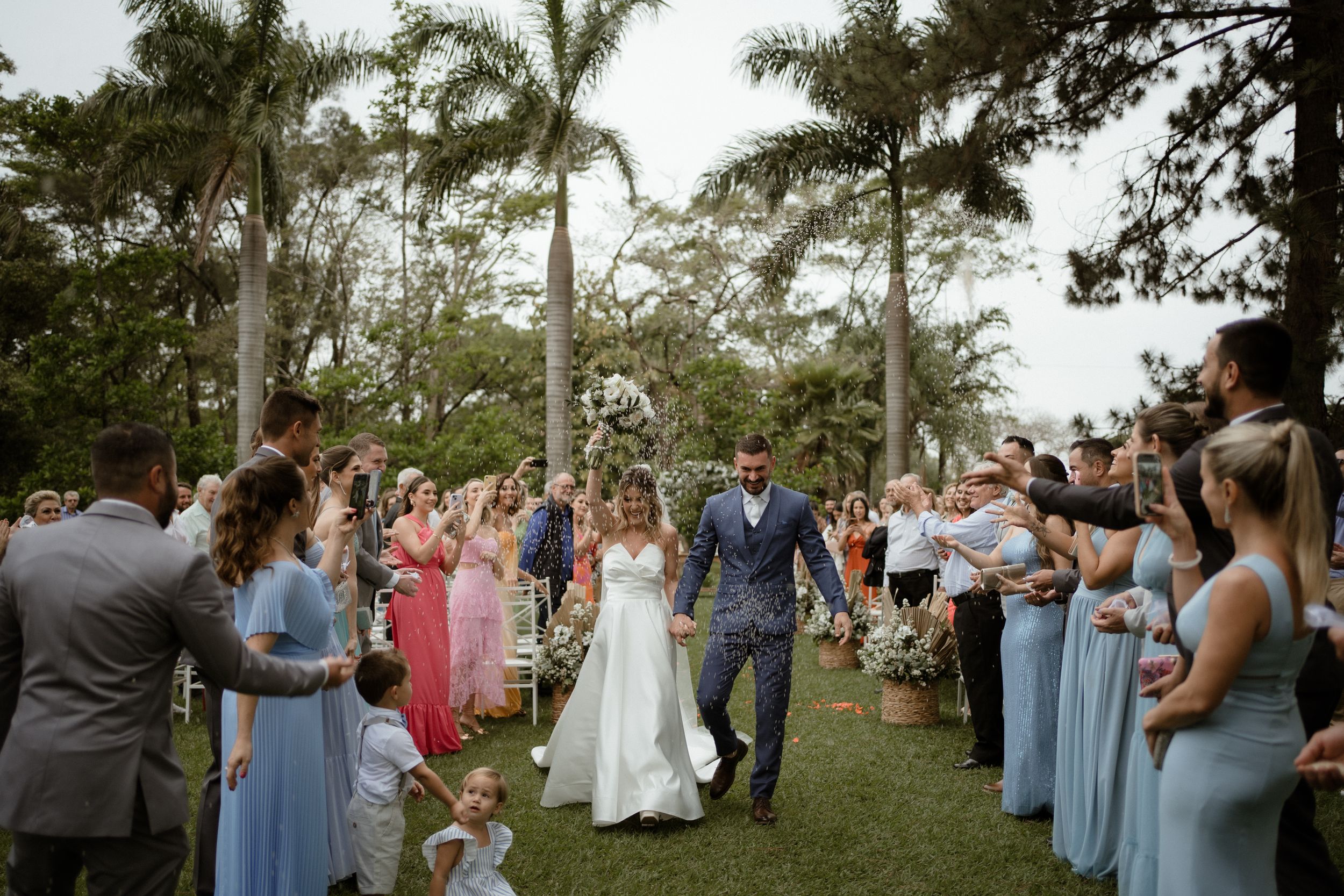 Wedding party exits ceremony through floral-lined aisle with guests throwing flower petals.
