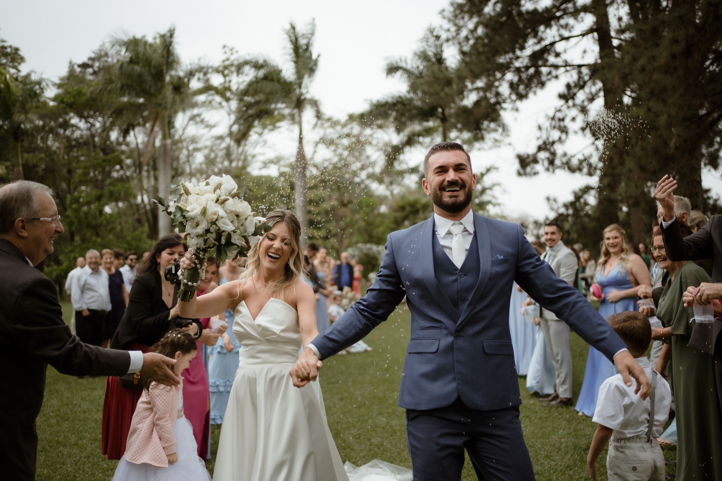 Joyful newlyweds celebrate with guests at outdoor wedding reception surrounded by palm trees.