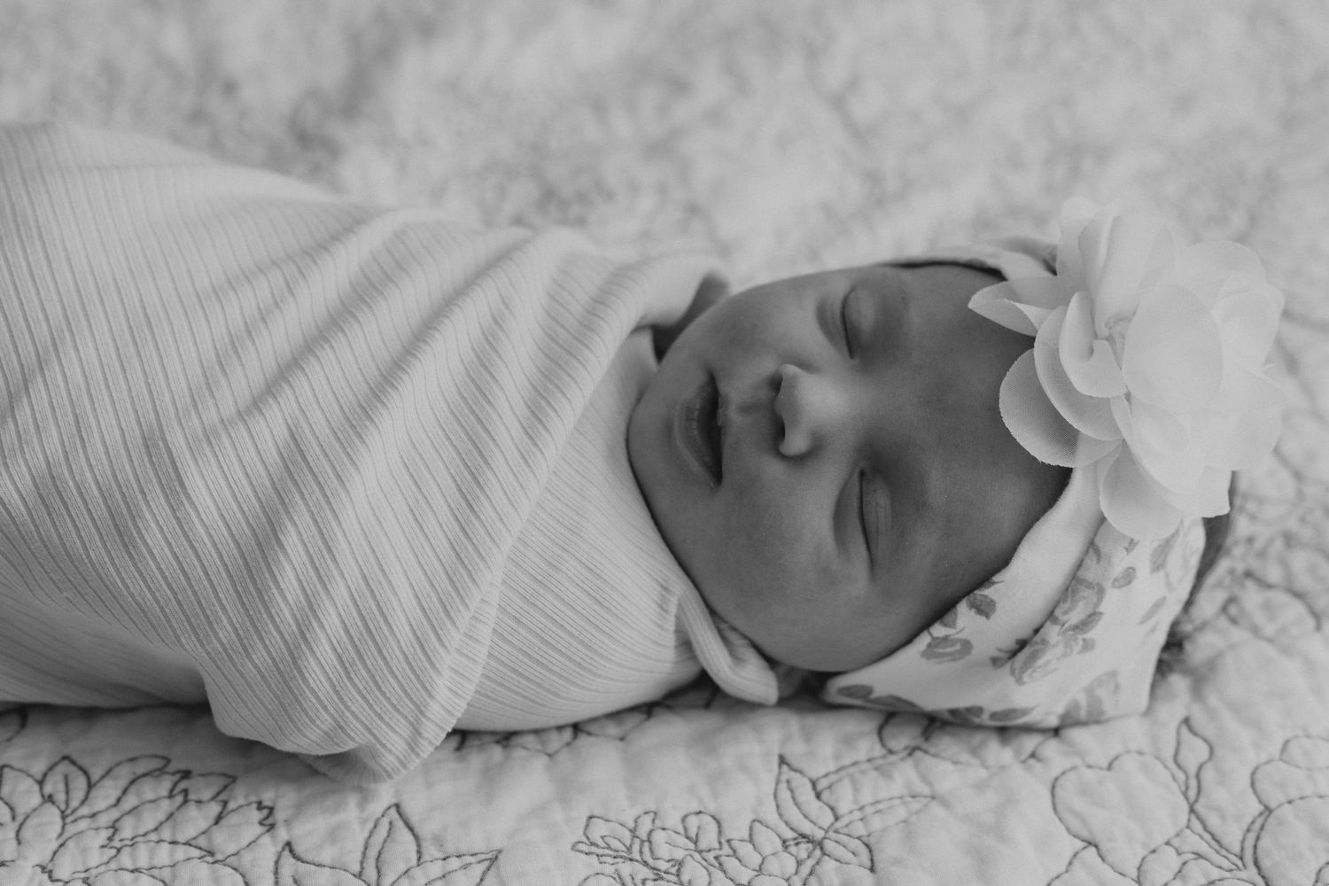 A newborn baby wrapped in white swaddle sleeps peacefully on textured bedding.