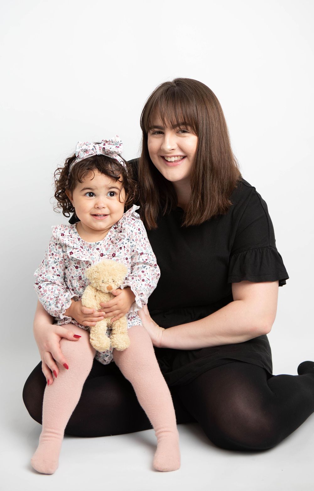 Smiling mother in black dress sitting with her curly-haired toddler daughter holding a teddy bear.