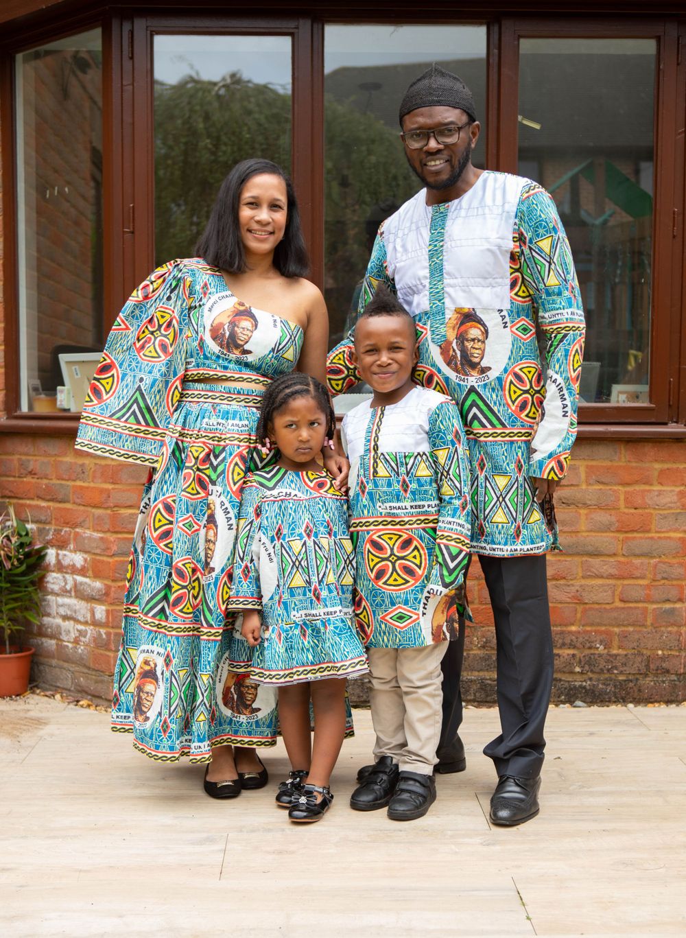 Family of four wearing matching vibrant African print outfits standing outside their home.