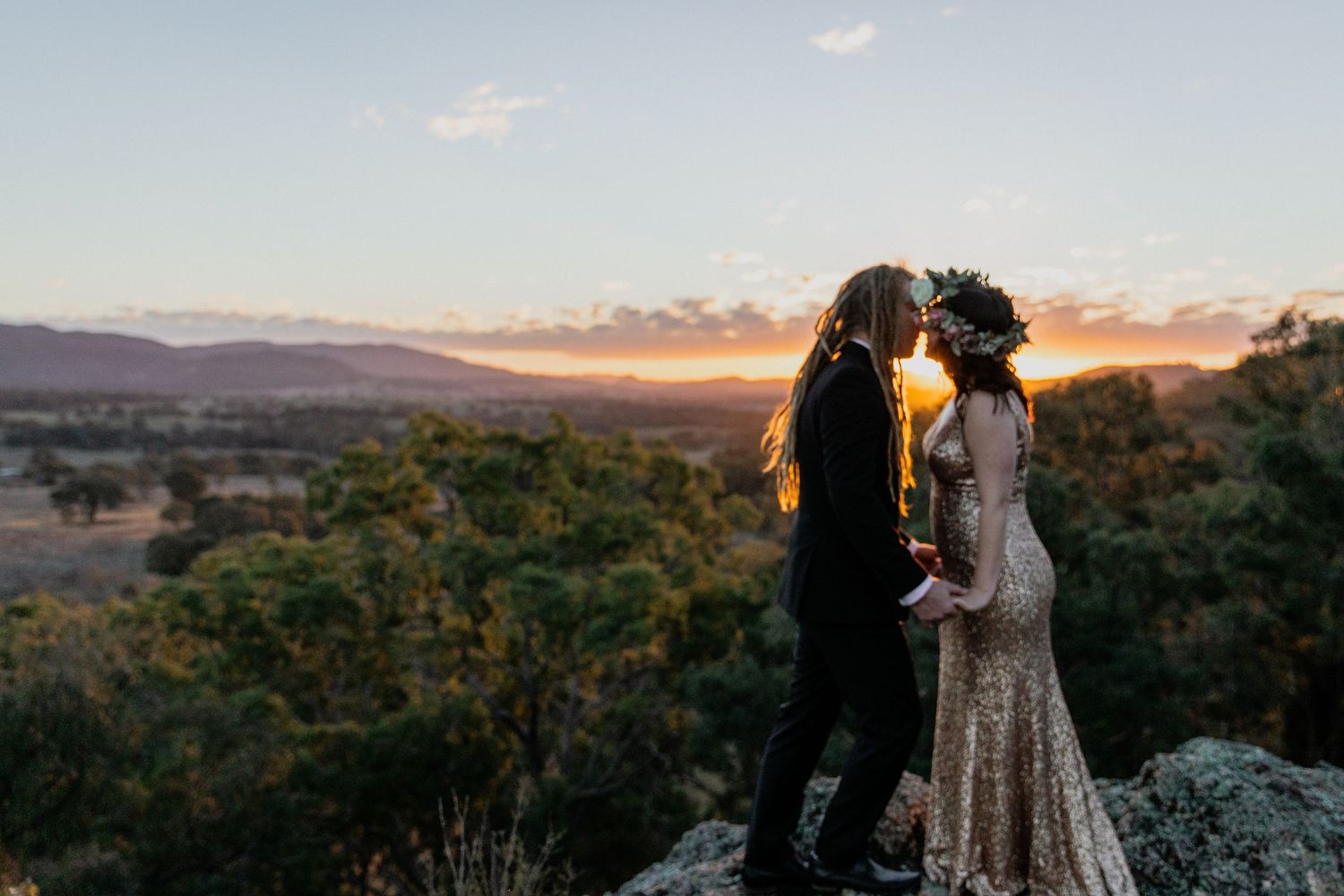 Couple sharing a sunset kiss on rocky outcrop, bride in gold gown with floral crown, groom in black, mountains behind.