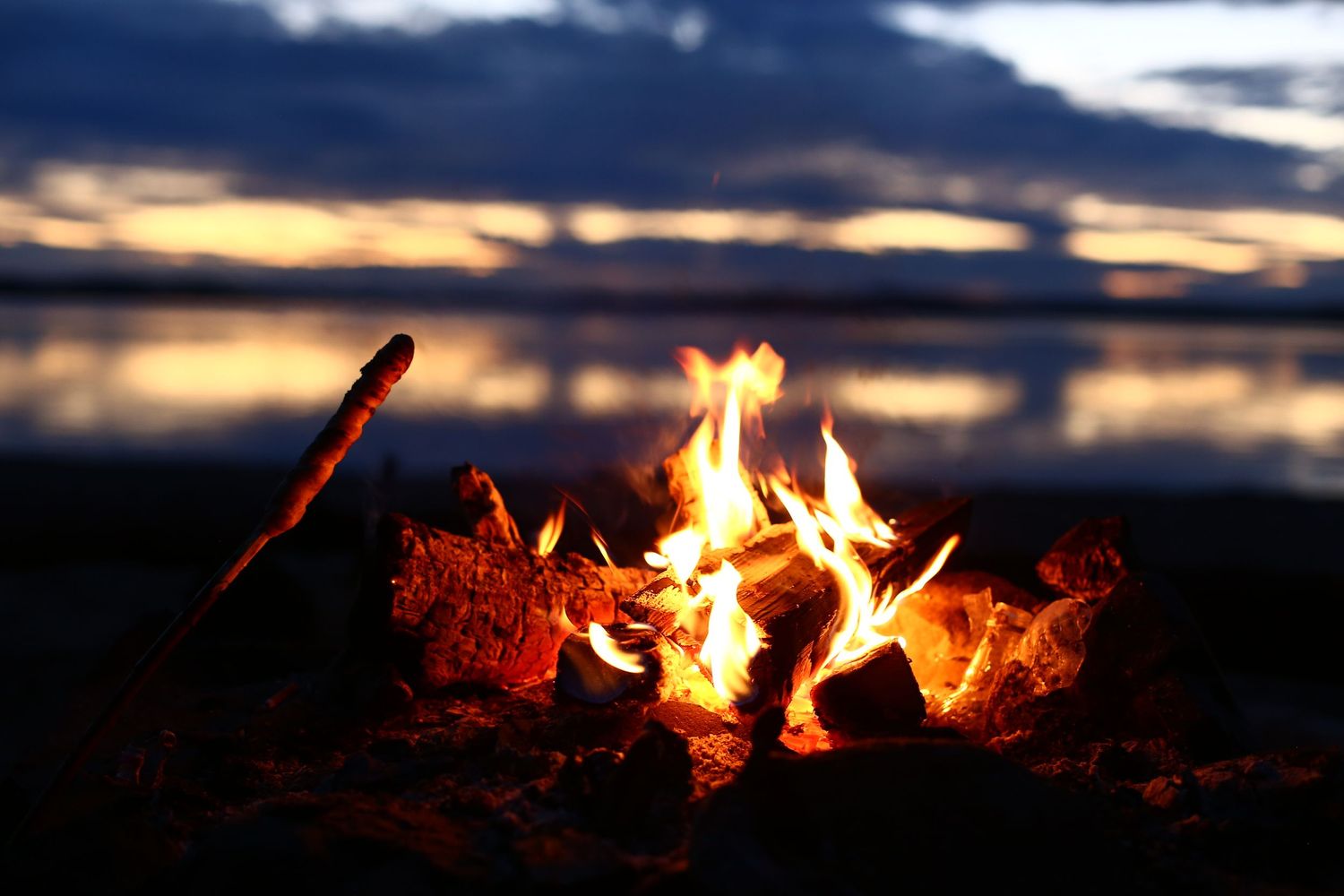 Crackling campfire burns brightly on a lakeshore at dusk with colorful sunset clouds in background.
