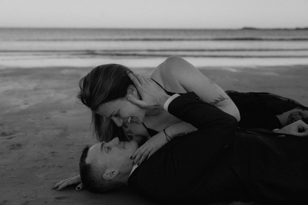 Romantic black and white photo of a couple embracing while lying on a sandy beach at twilight in Maine