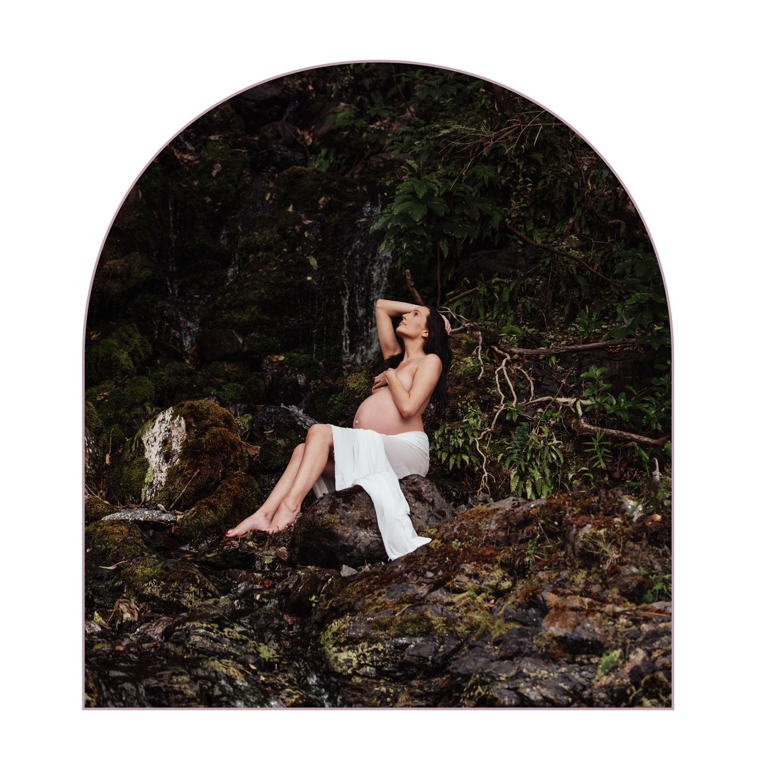 A dreamy maternity portrait of figure in white dress sitting on mossy rocks in a dark forest setting.