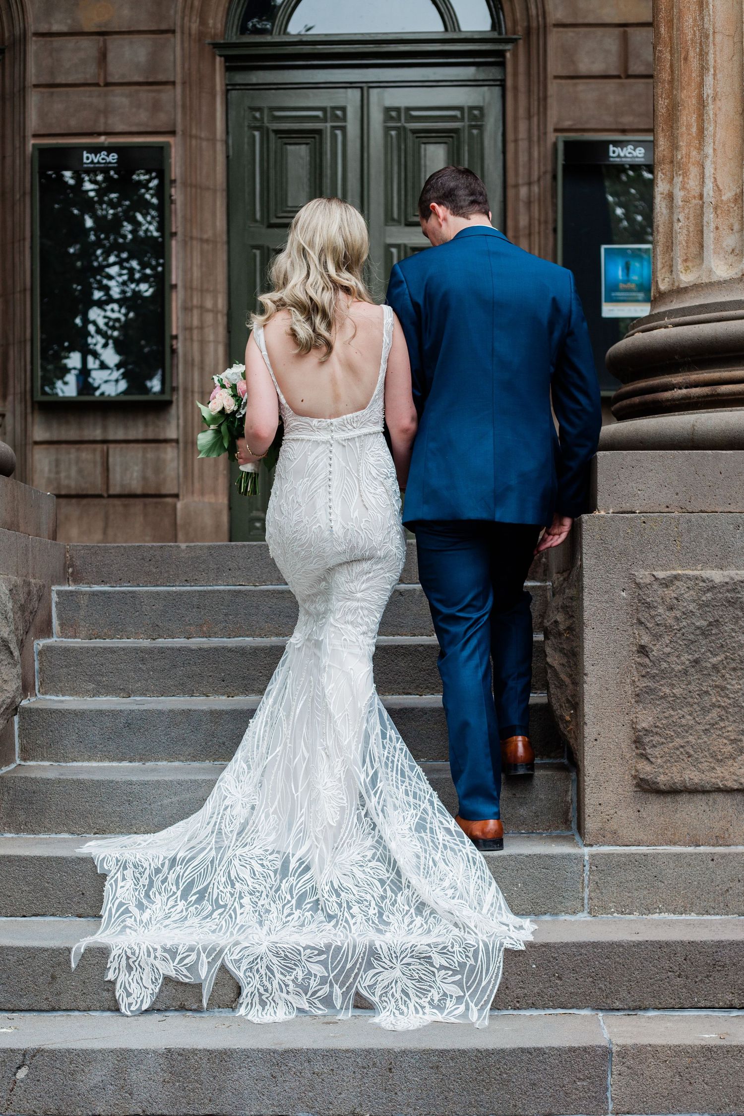 A couple in wedding attire ascends stone steps towards an ornate wooden door.