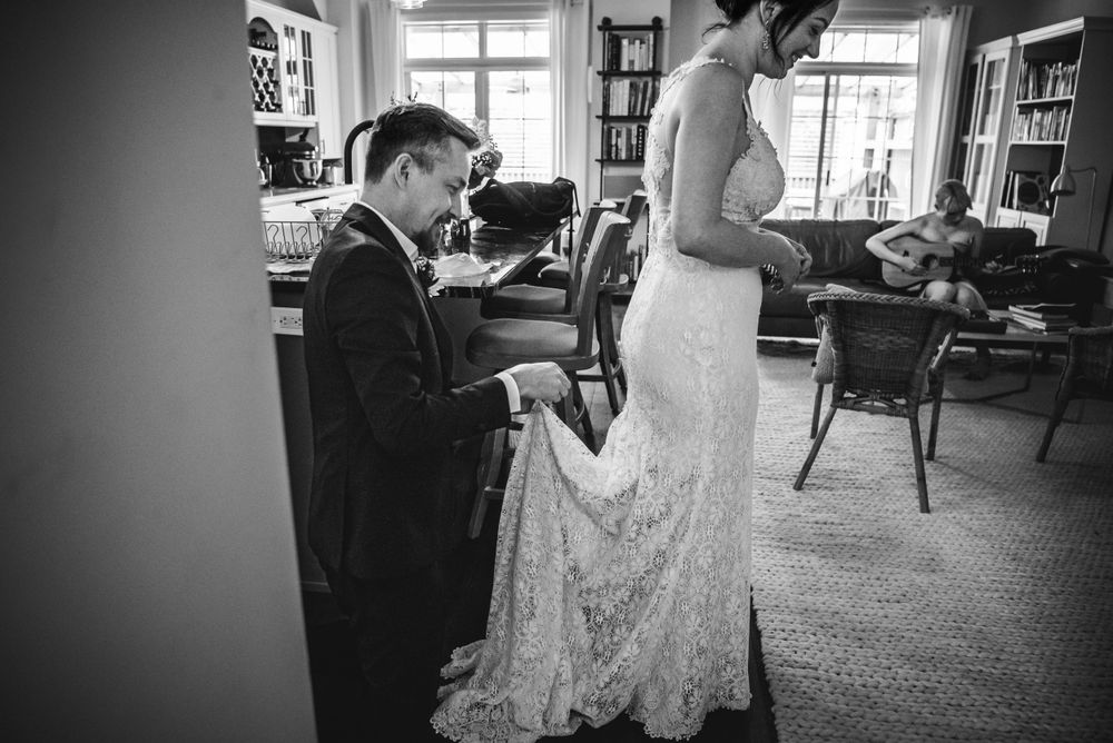 Black and white image of a man kneeling behind his soon to be bride and helping her fix the back of her dresses train.