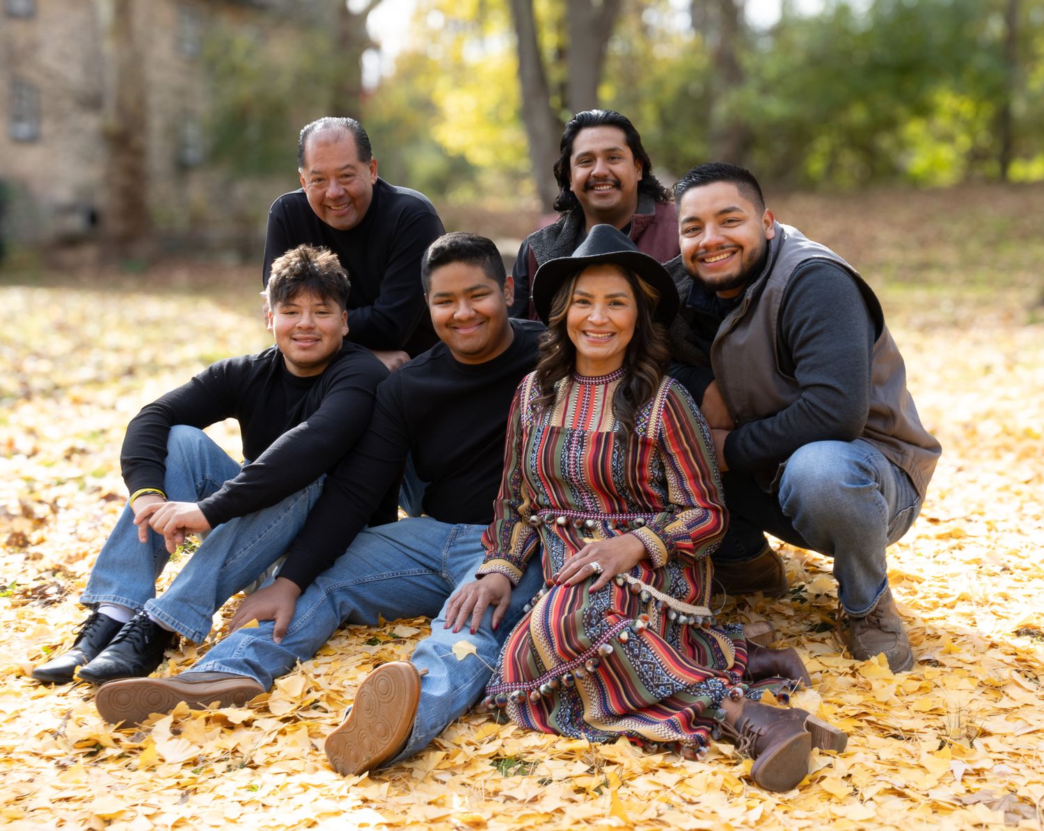 Joyful Latino family of six posing on golden autumn leaves in a park, with a woman in a colorful dress at center.