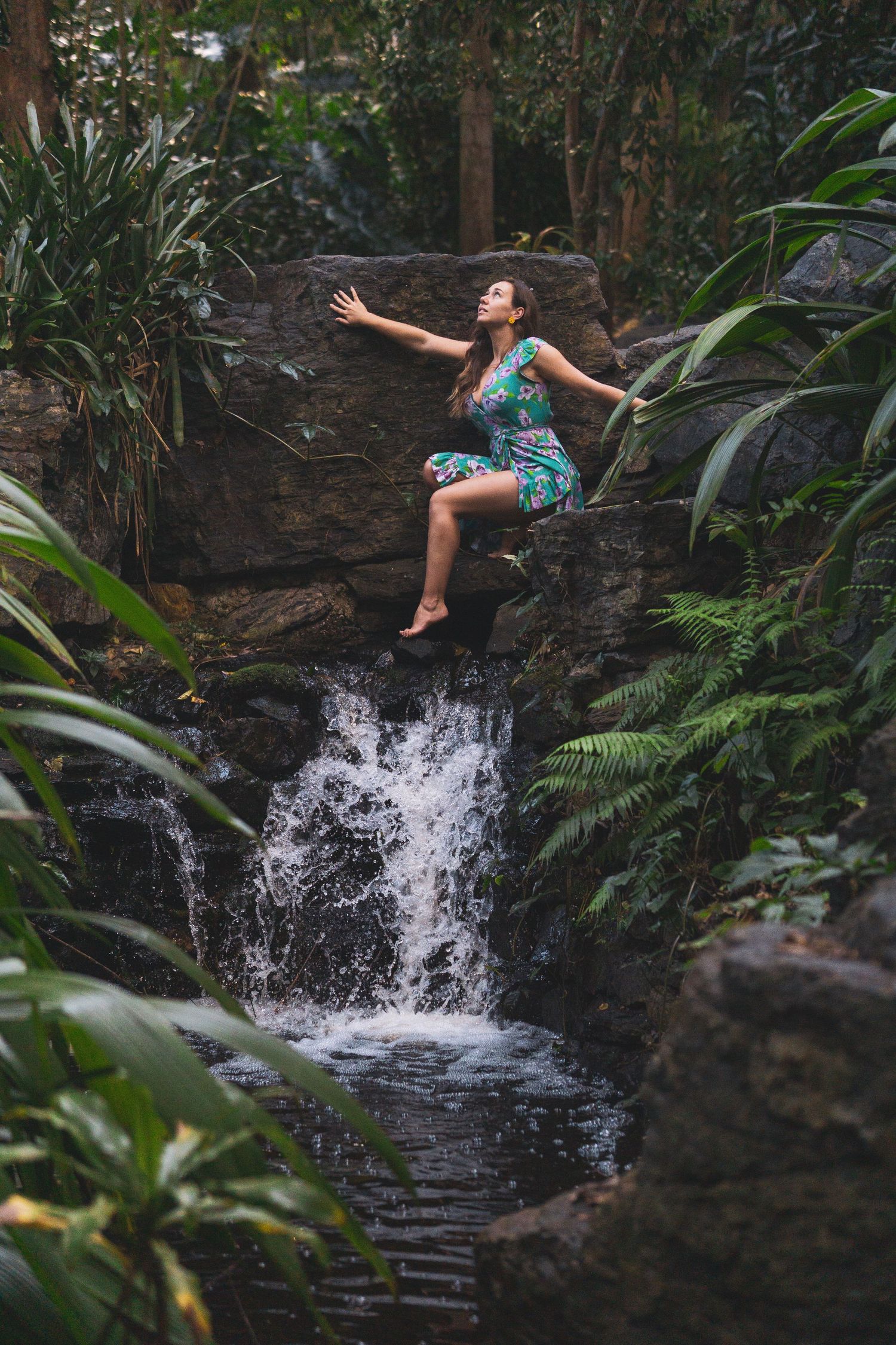 Person in summer dress leaps joyfully near a cascading waterfall surrounded by jungle foliage.