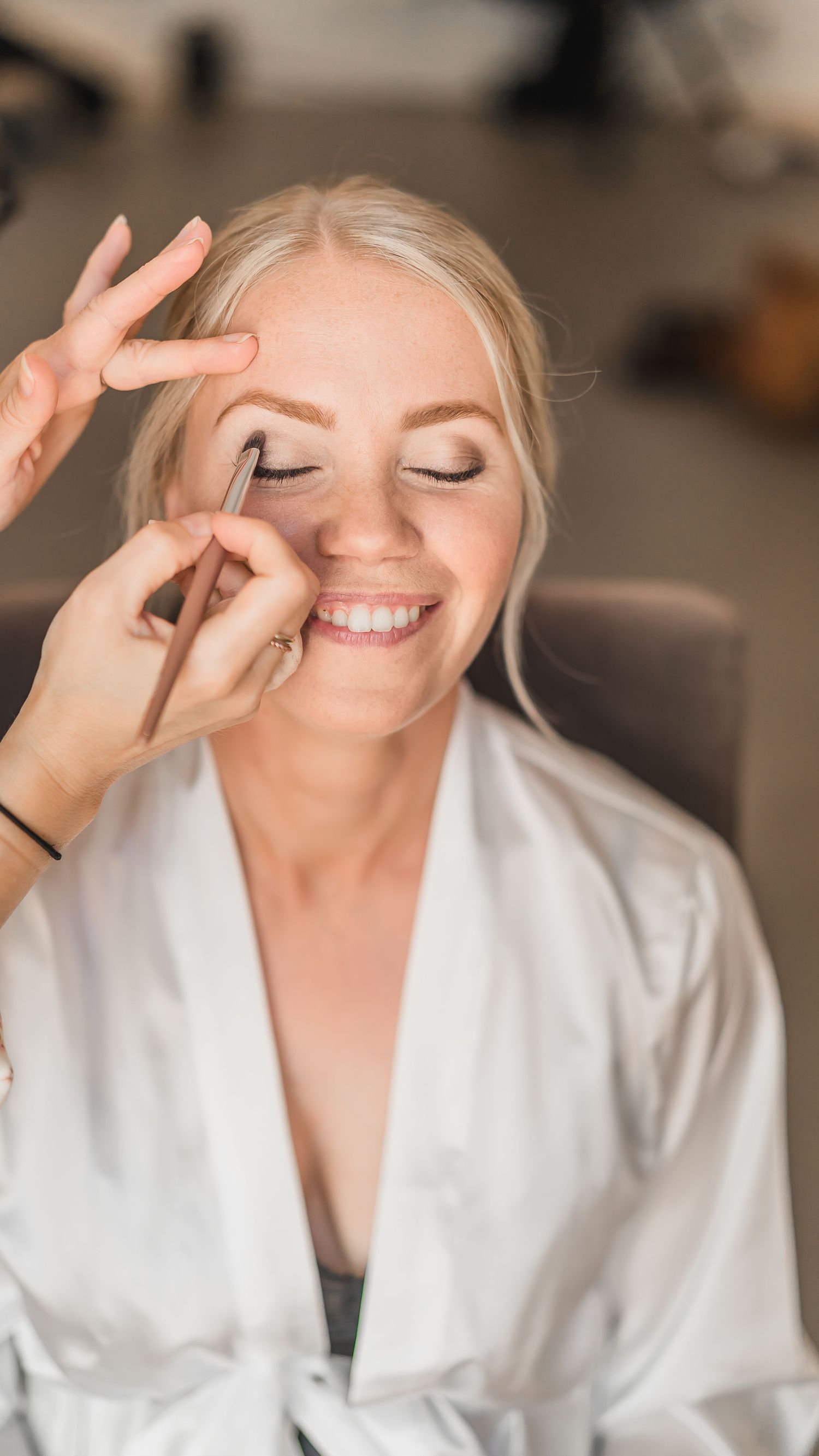 Makeup artist applying eyeliner to a smiling blonde bride in a white satin robe during bridal prep.