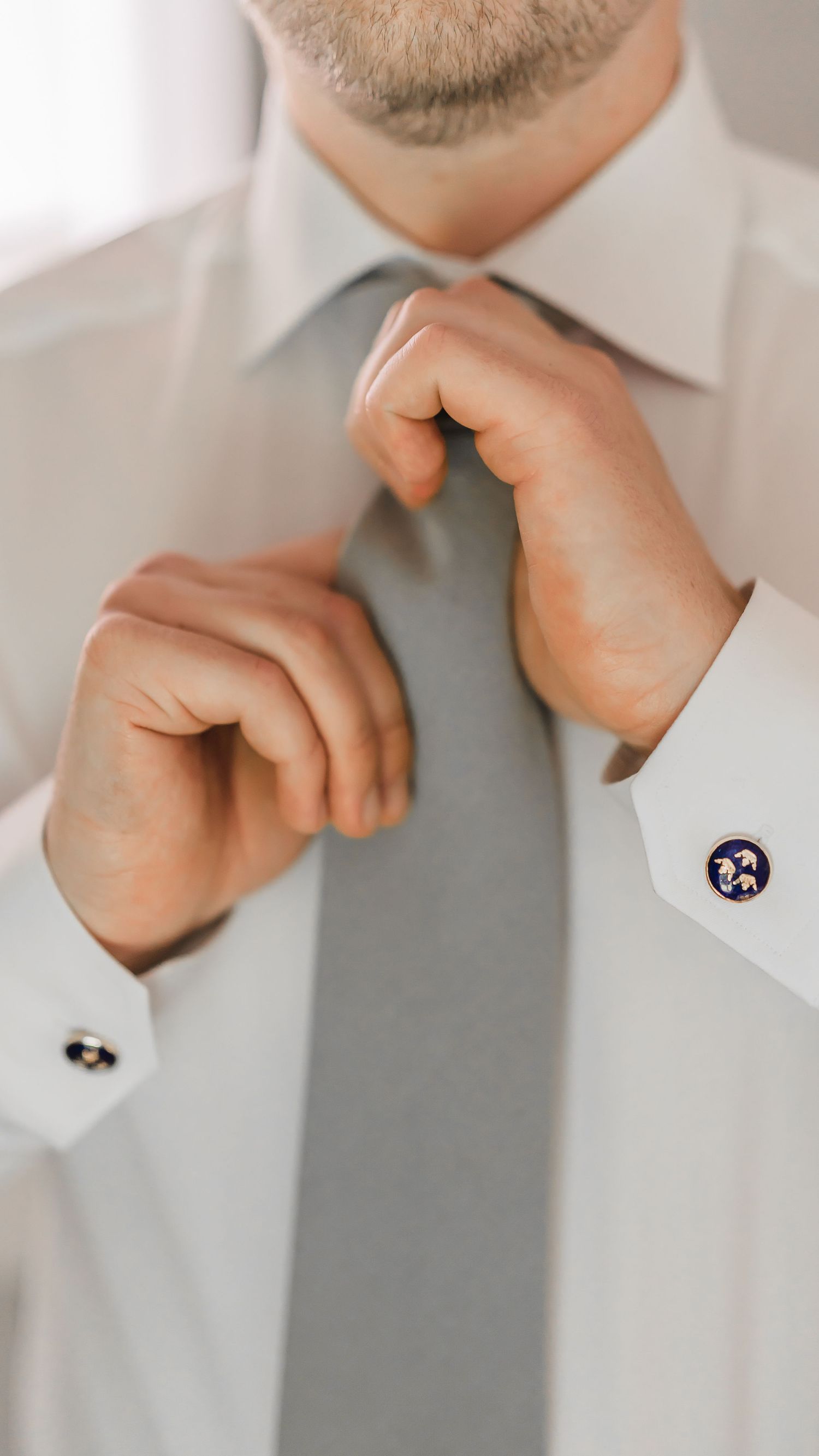 Close-up of groom adjusting gray tie wearing white dress shirt with decorative blue cufflinks.