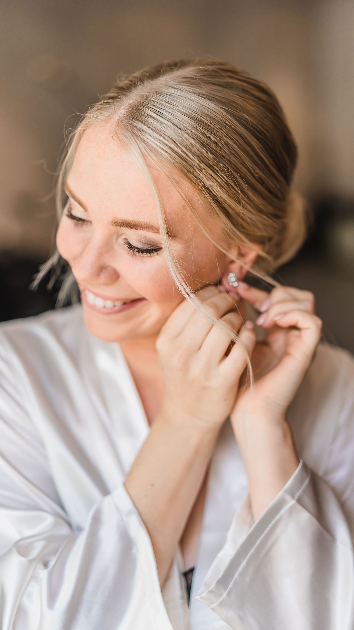 Smiling blonde bride in white robe putting on delicate pink earrings while getting ready.