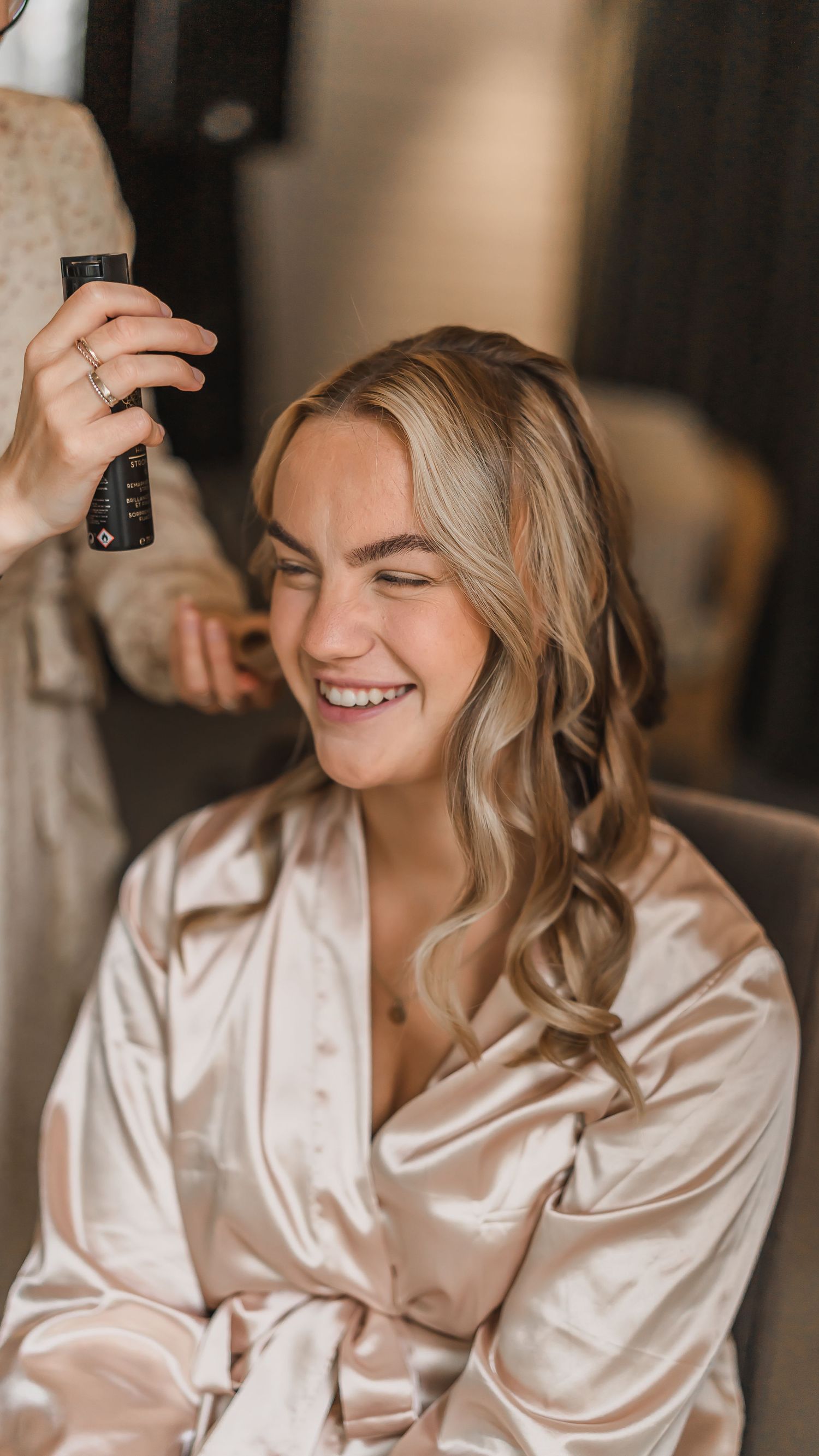 Blonde bride laughing joyfully in a satin robe while a stylist curls her hair before the wedding.