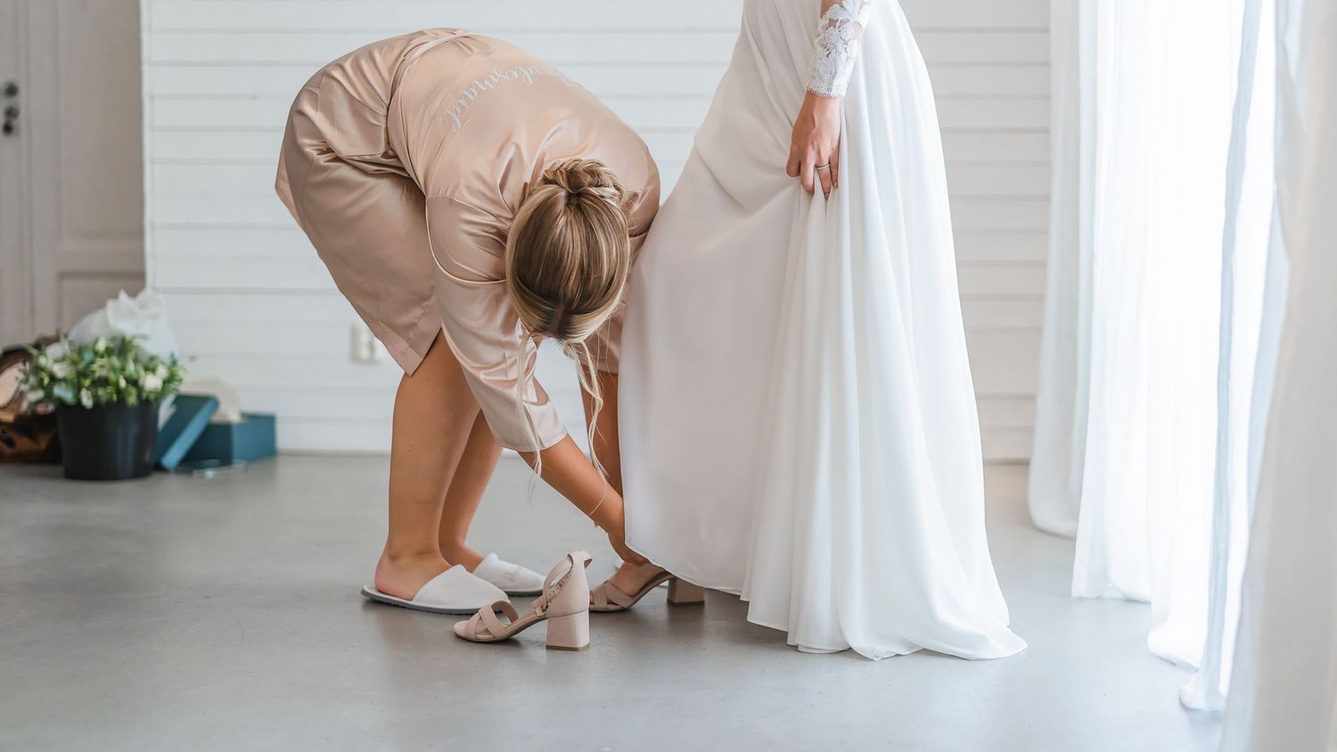 Bridesmaid kneeling to help bride put on white wedding shoes beneath flowing bridal gown.