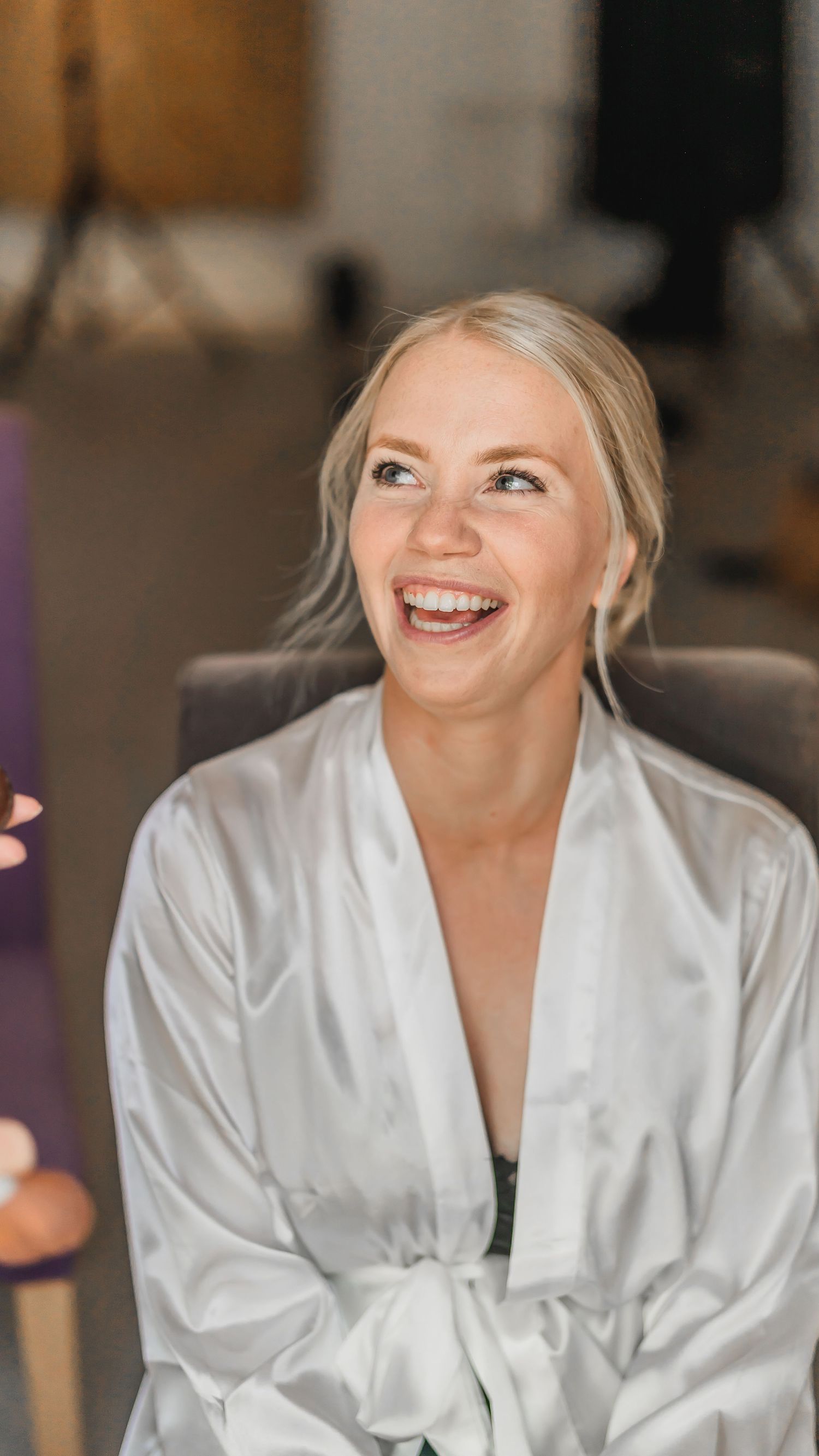 Happy blonde bride laughing joyfully in a white satin robe during bridal hair and makeup preparations.