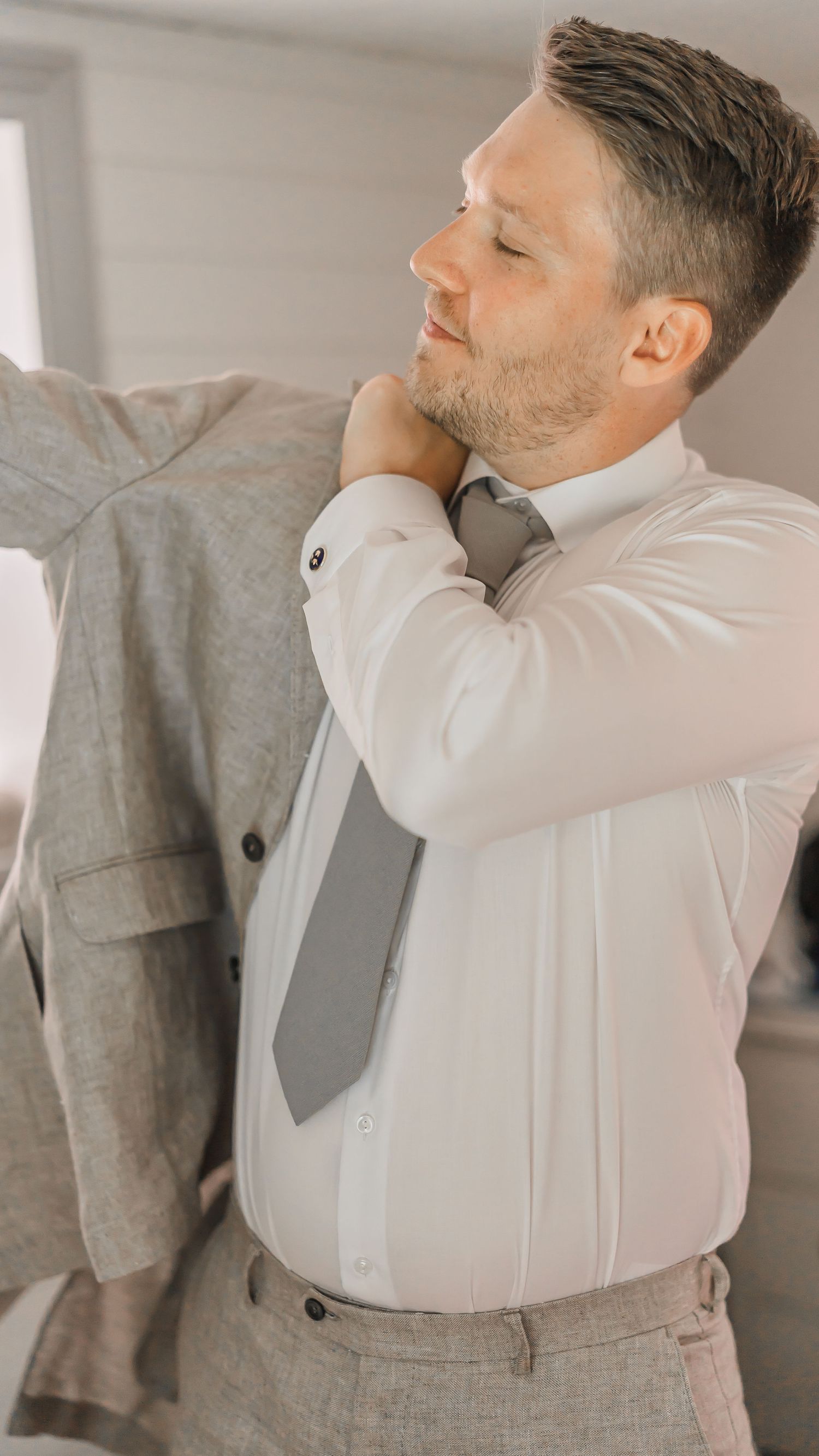 Groom smiling while putting on his light gray linen wedding suit jacket before the ceremony.