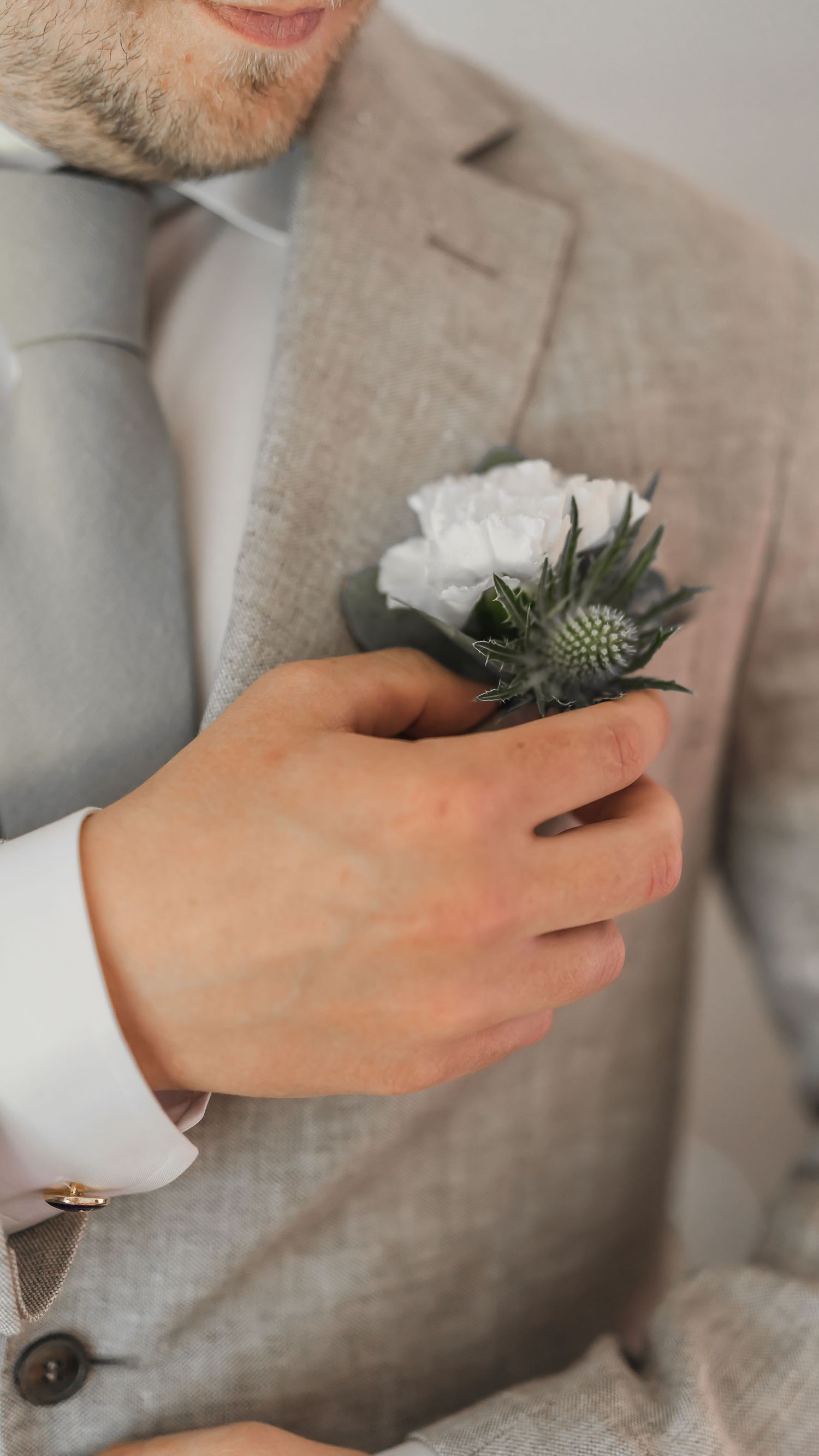 Groom holding a white carnation and thistle boutonniere against his gray linen wedding suit jacket.