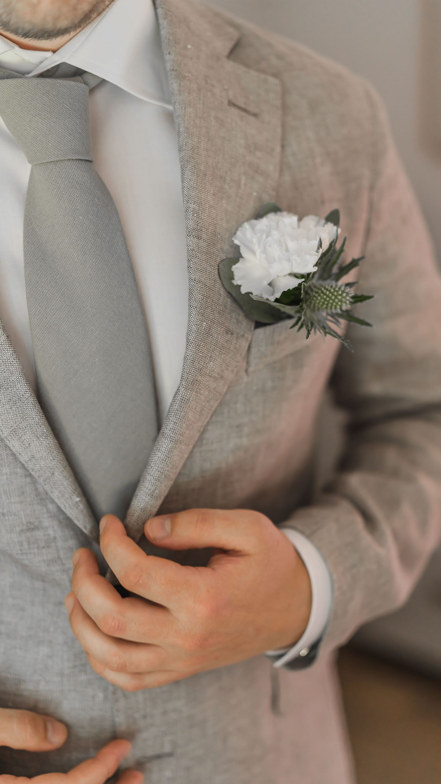 Close-up of groom in beige linen suit adjusting jacket with white carnation and thistle boutonniere.
