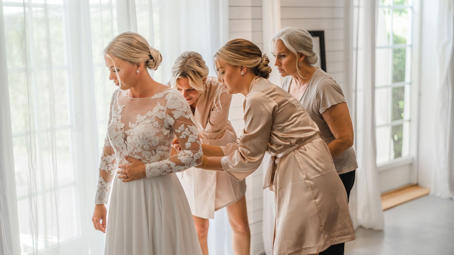 Bridesmaids helping blonde bride into her elegant white lace wedding dress before ceremony.