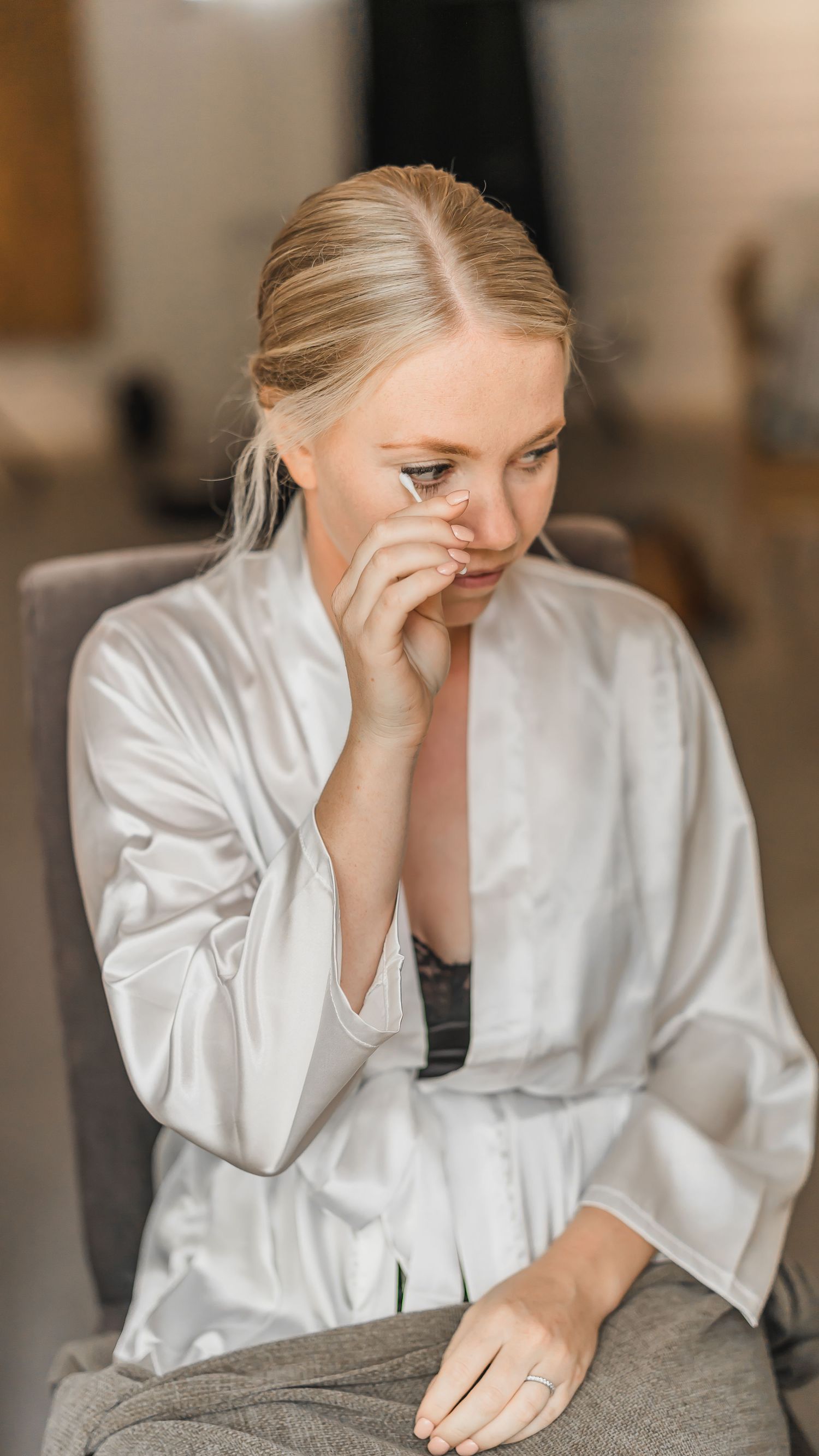 Emotional bride wiping tears while sitting in a chair wearing a white satin robe during wedding preparations.