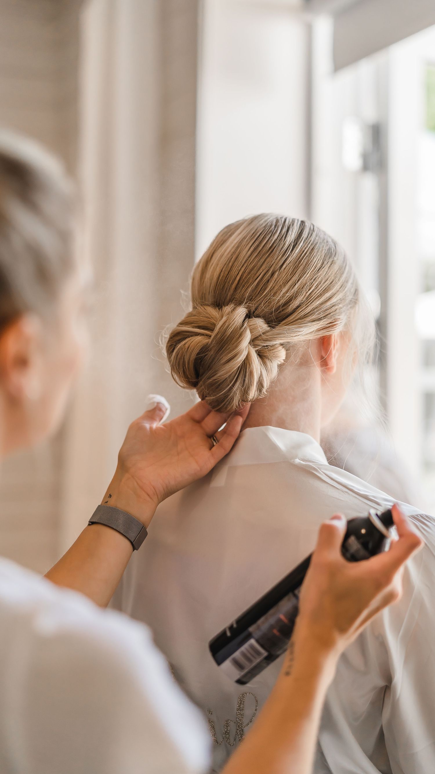 Hairstylist spraying and finishing an elegant bridal updo on a blonde bride in a white robe.