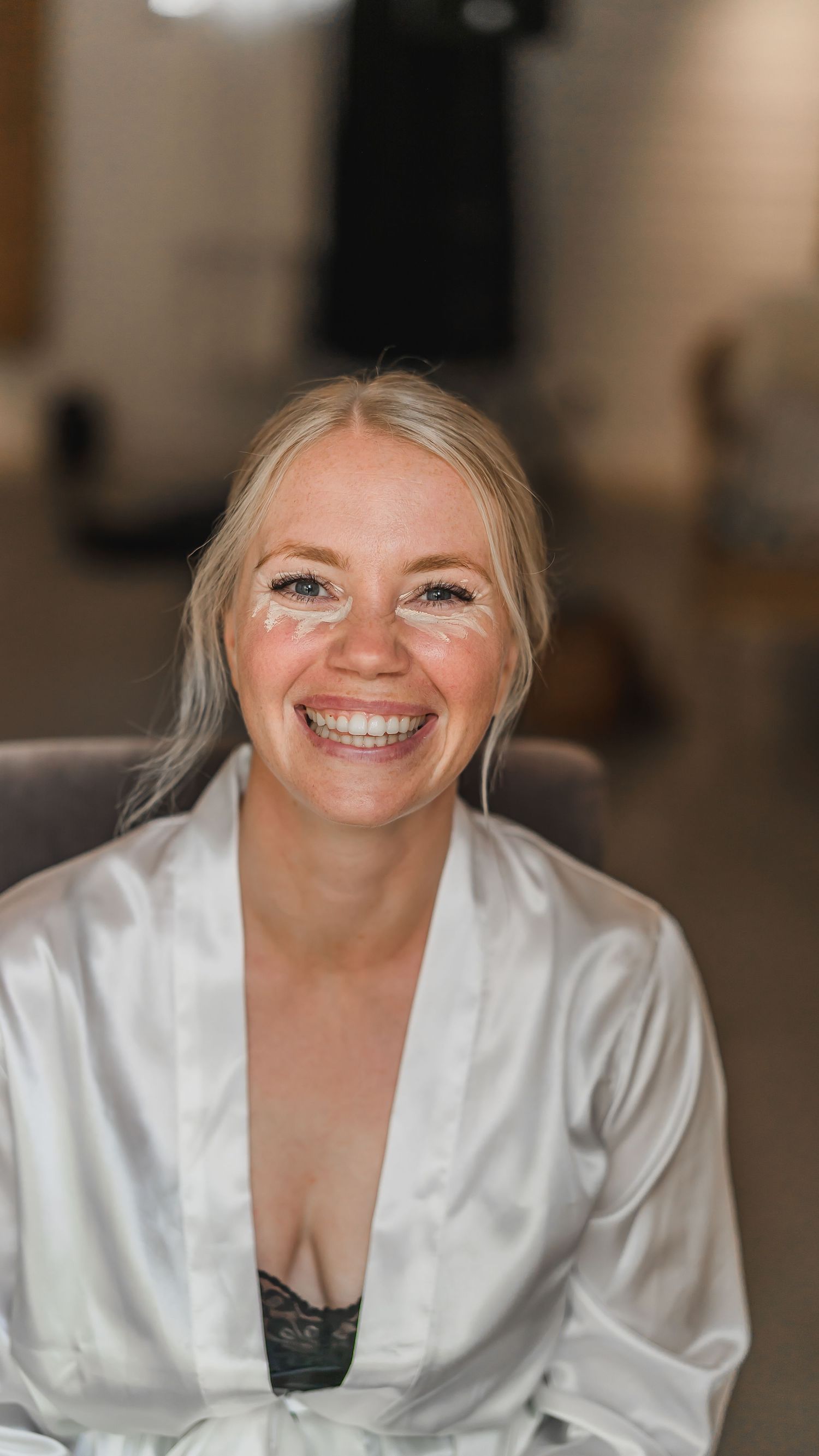 Smiling blonde bride in a white satin robe with under-eye patches during wedding morning preparations.