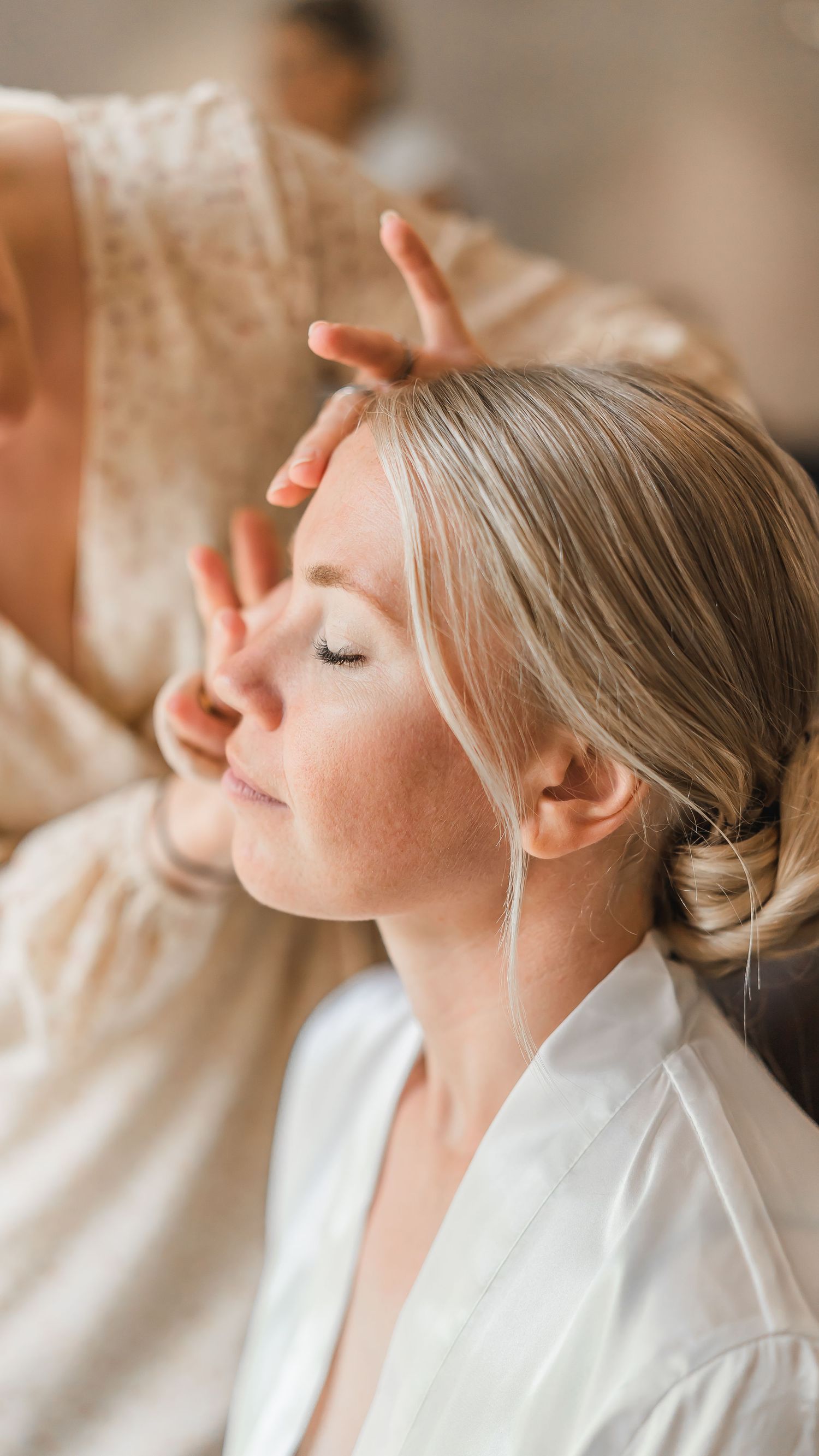 Makeup artist carefully applying bridal makeup to a serene blonde bride in a white robe before wedding.