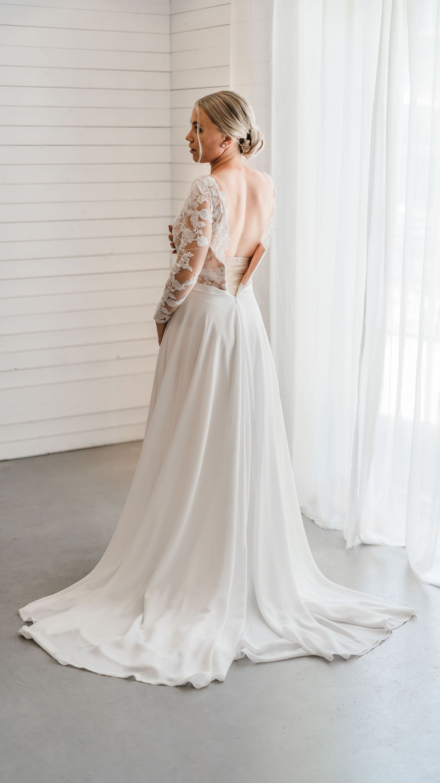 Bride in stunning backless lace wedding gown with flowing skirt standing by sheer curtains.