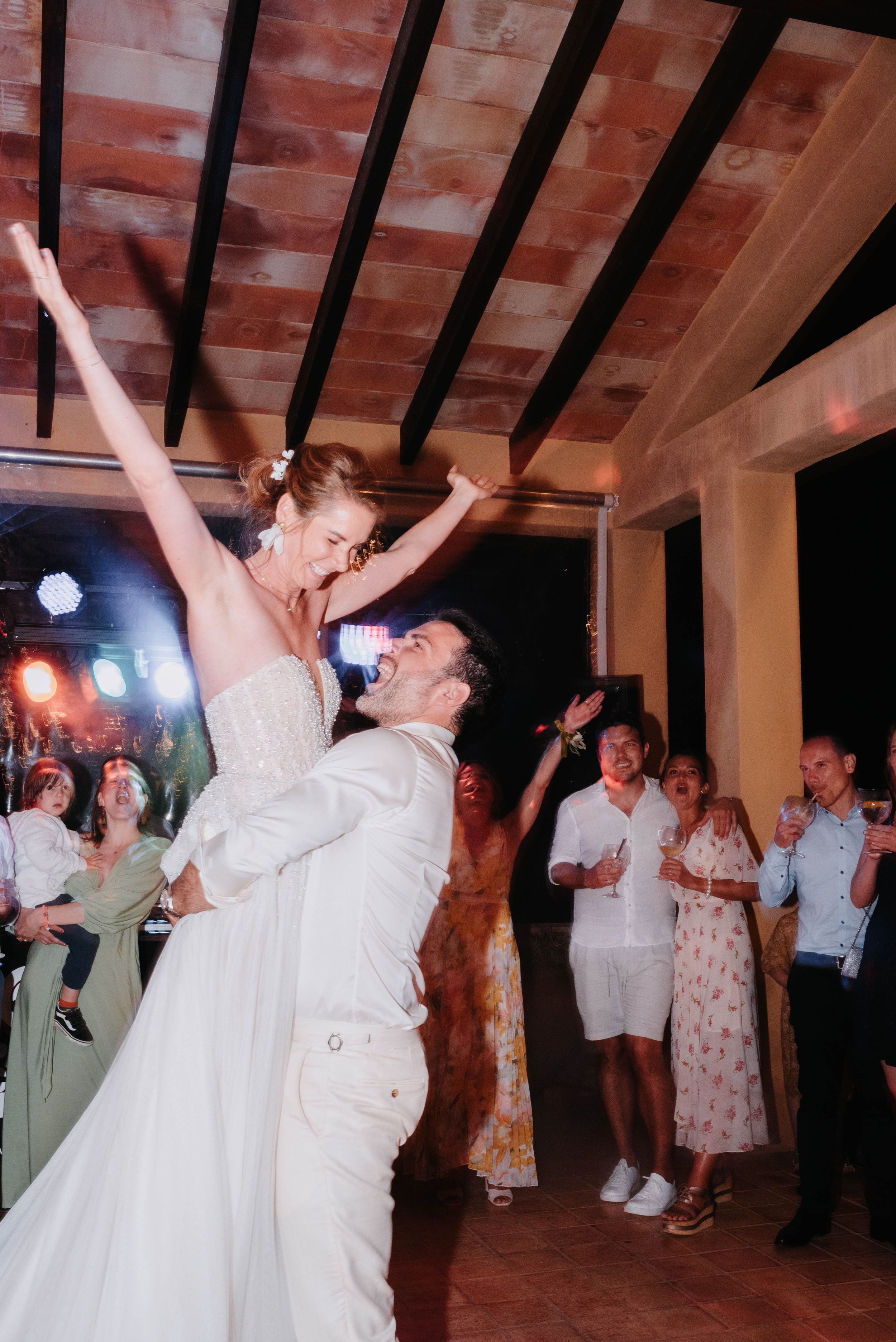 Celebratory wedding dance lift captured under rustic wooden beams with party lights.