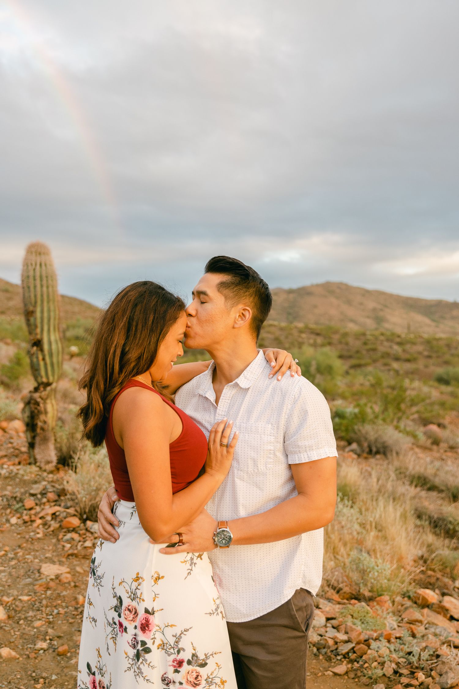 A couple stands in front of a rainbow. The man is kissing the woman's forehead and there's desert and cactus behind them. The sun is setting