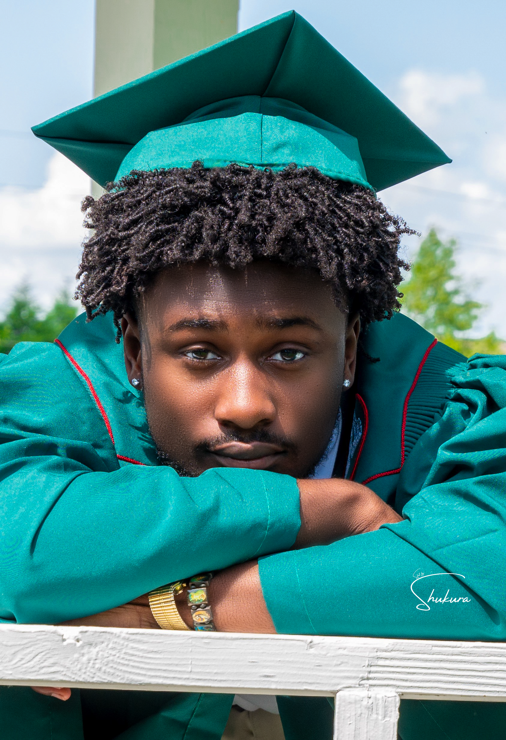 A graduate wearing a teal graduation cap and gown rests their head on a white railing during a graduation photoshoot outdoors.