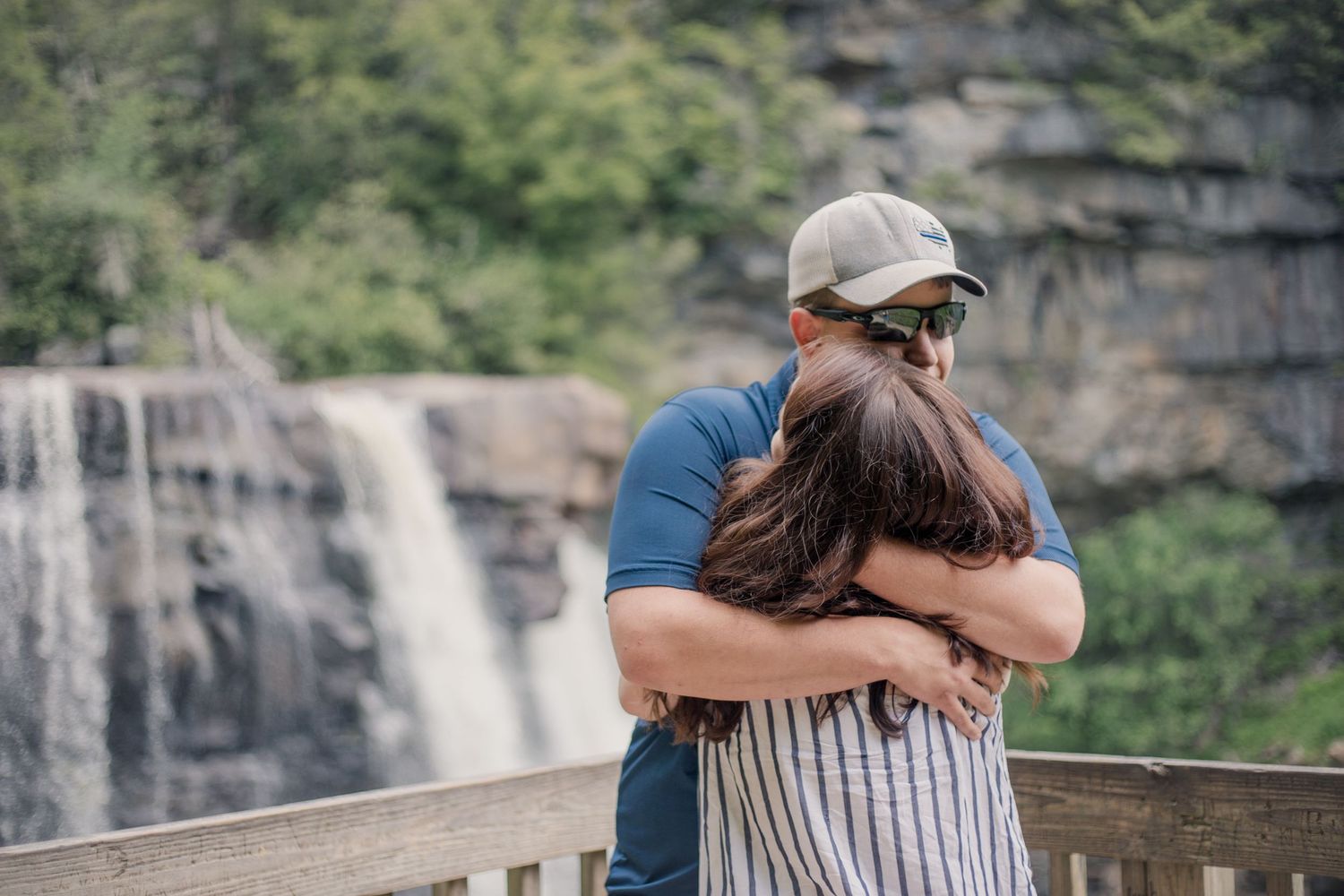 Two people sharing an emotional moment on a wooden deck with dramatic cliffs and waterfalls behind them.