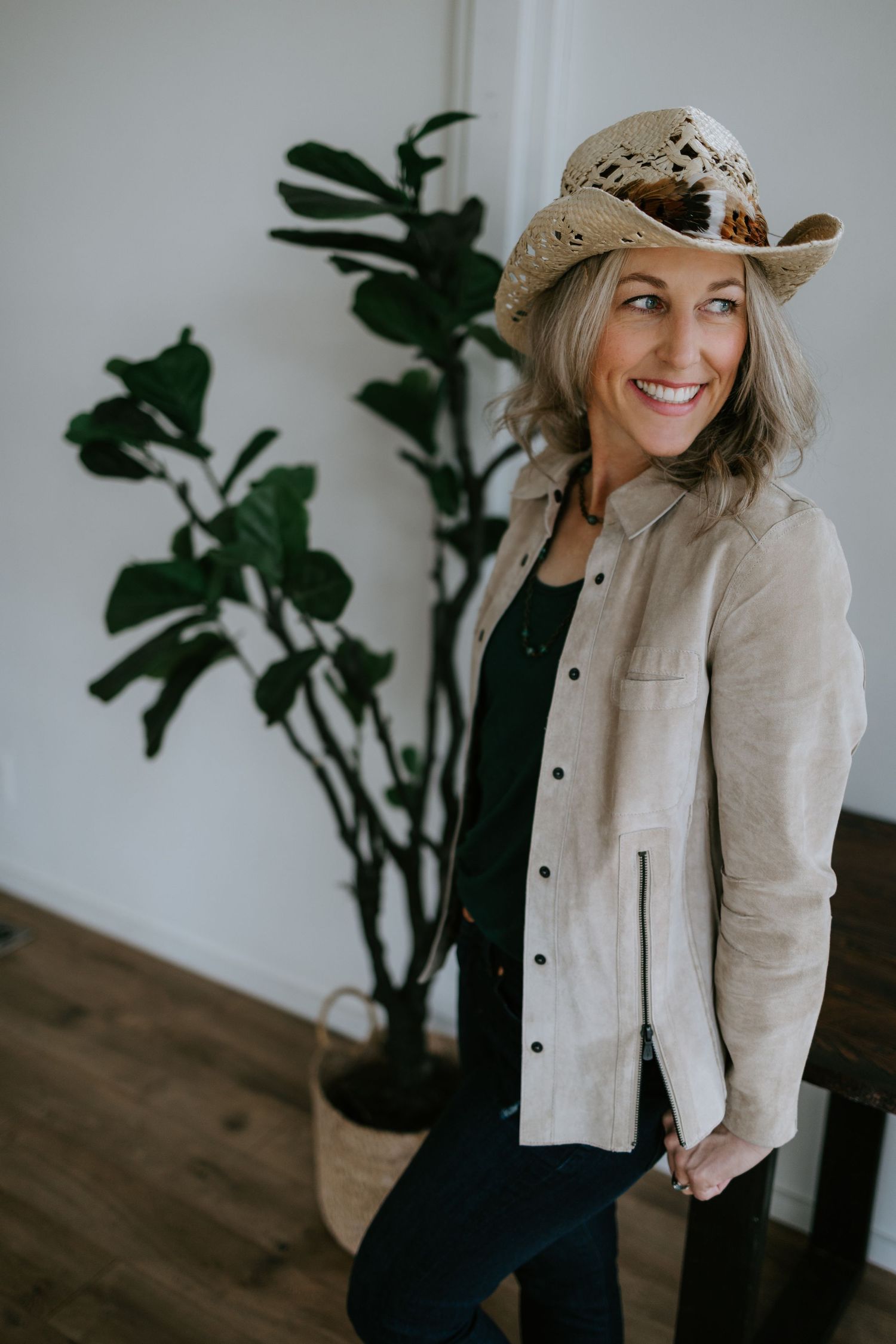 Casual western-inspired outfit with straw hat and beige jacket next to indoor plant against white wall.