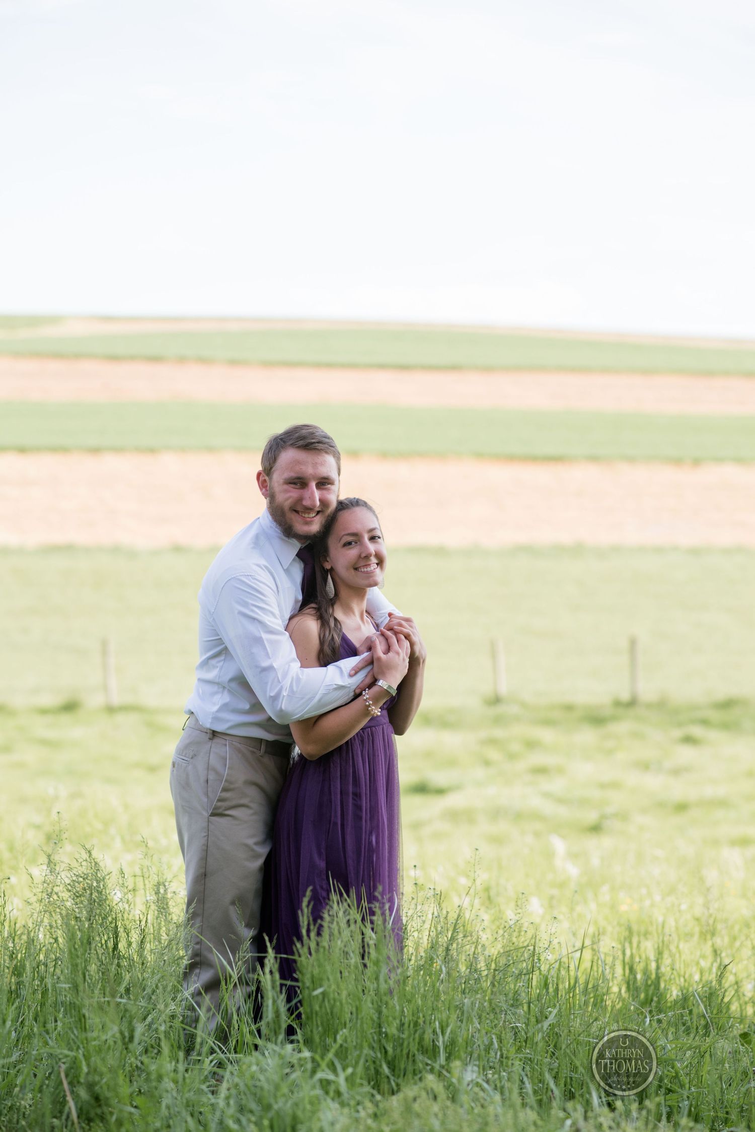 Couple embrace in a rural field setting with rolling hills in the background.