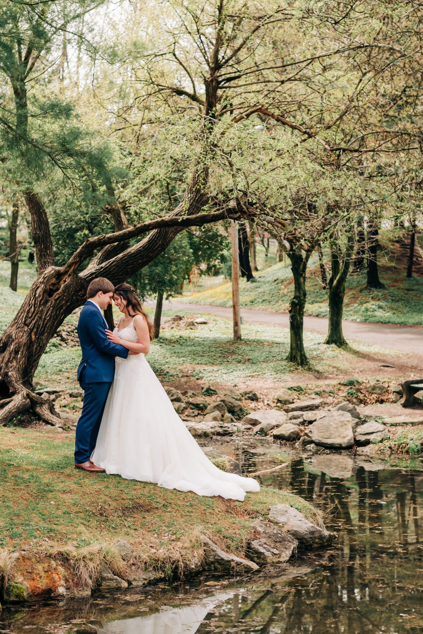 bride and groom pose for a portrait photo under a tree by a small creek