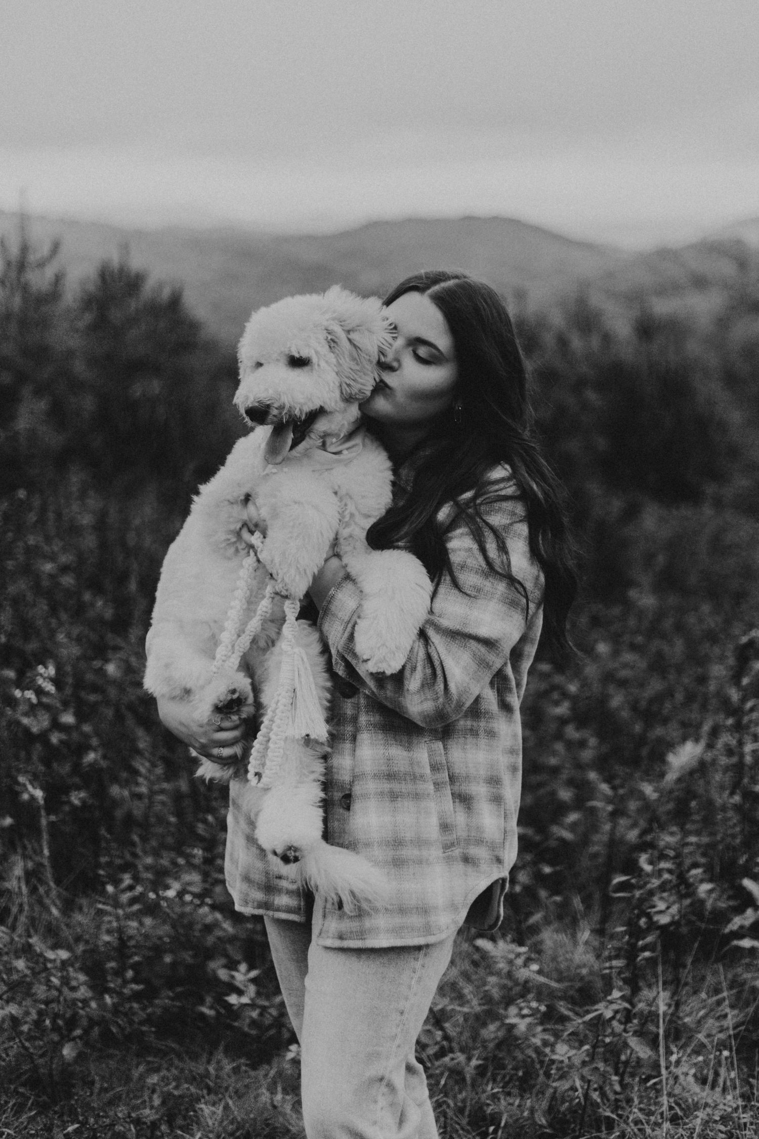 A person in plaid holding and kissing a fluffy white dog against a mountainous background.