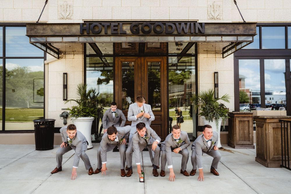Wedding party in grey suits poses playfully outside Hotel Goodwin entrance.