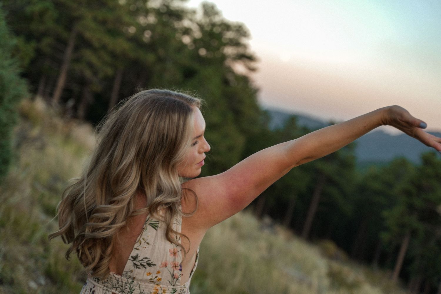 Person in floral dress stretches arm gracefully against mountain sunset backdrop.