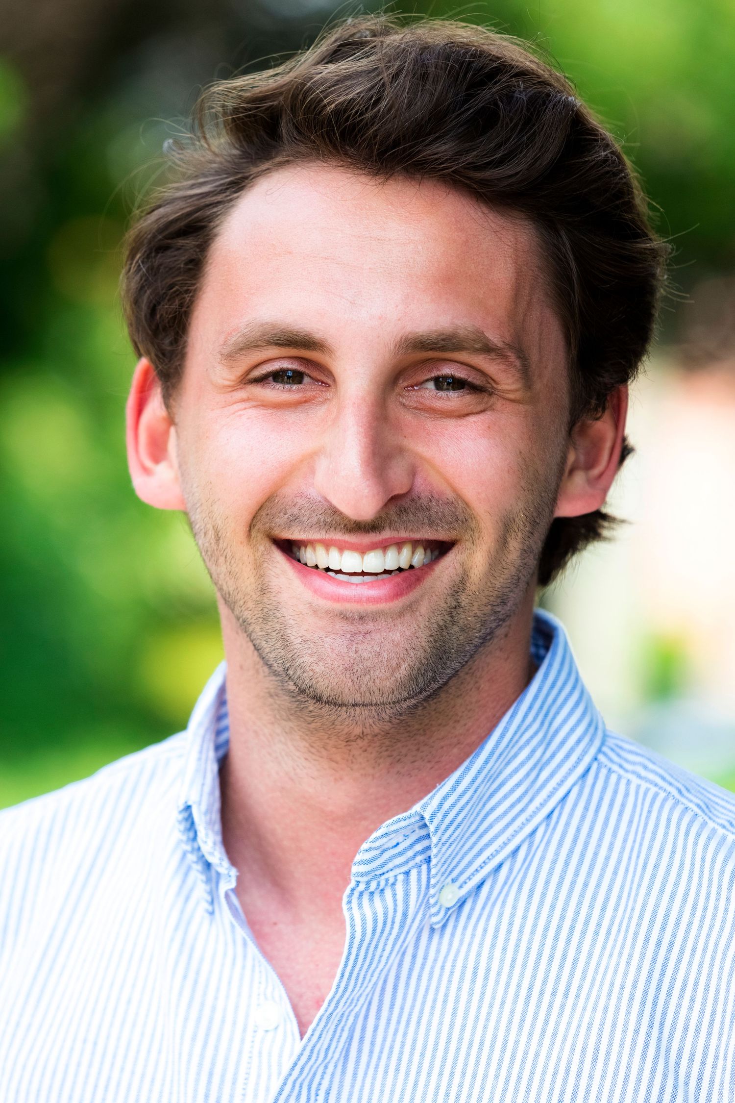 Professional portrait of person in striped button-down shirt with natural outdoor backdrop.