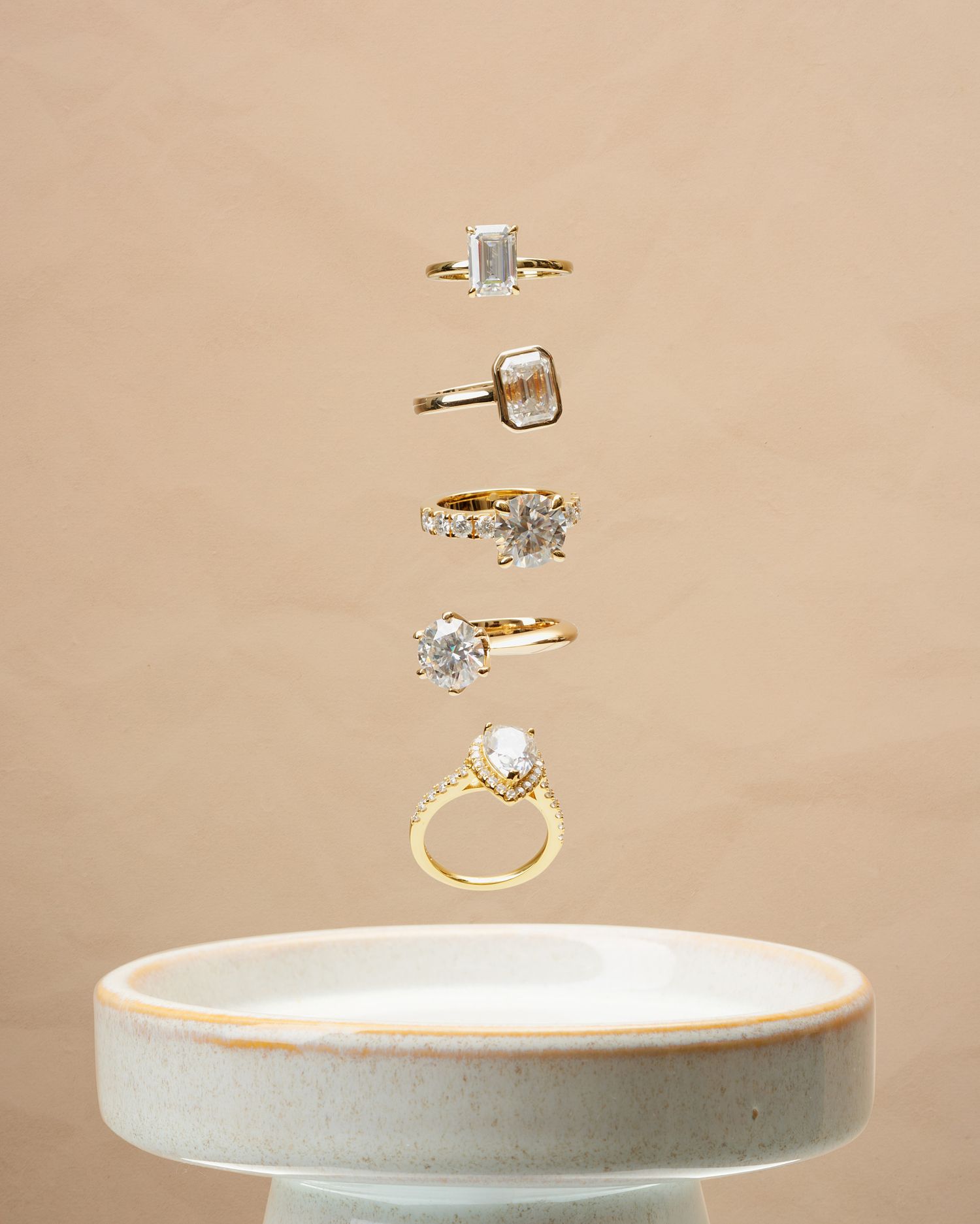 Five floating diamond engagement rings arranged vertically above a ceramic dish.