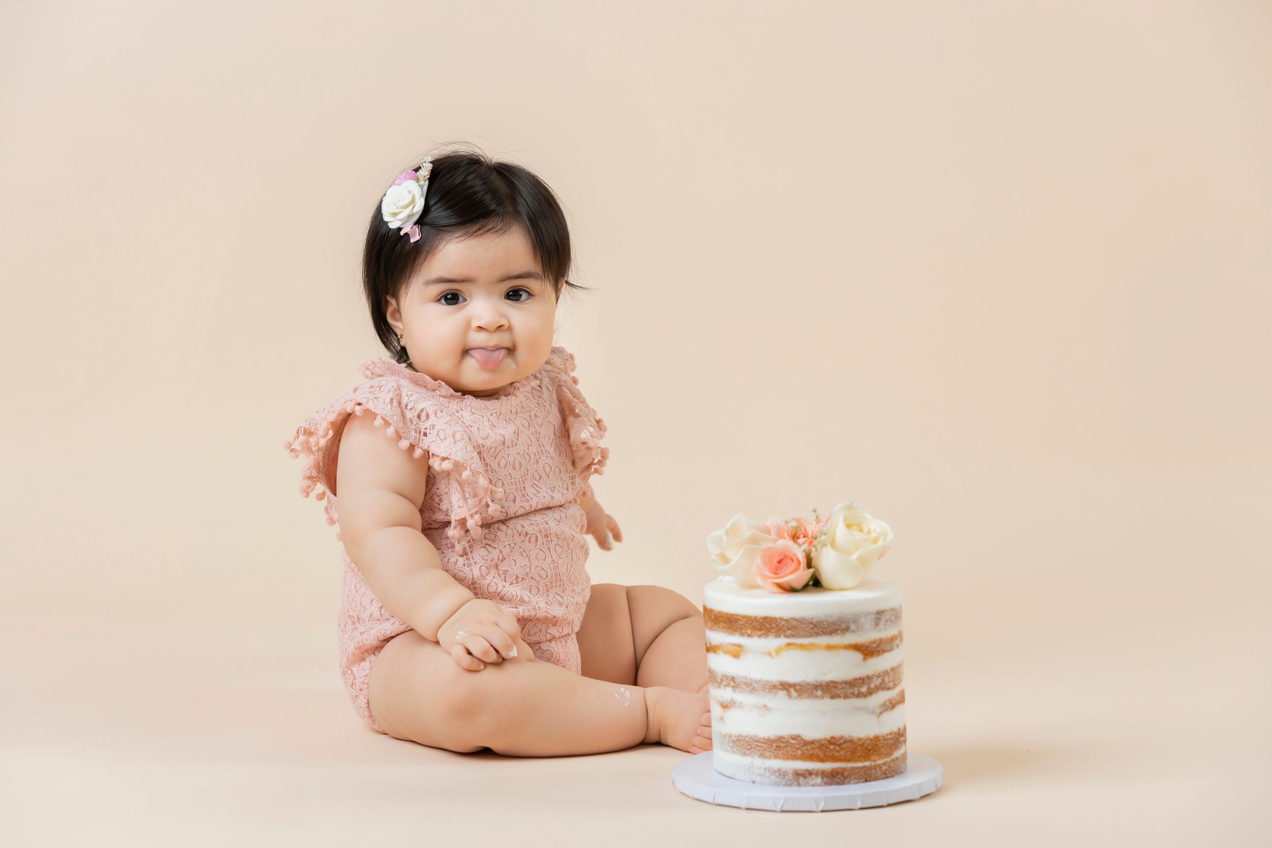 A baby in pink romper sits near decorated naked cake on pedestal against neutral background.