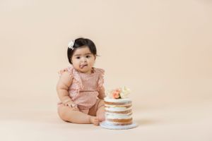 A baby wearing pink ruffled outfit poses next to tiered naked cake against beige backdrop.