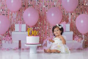 Pink birthday setup with white cake, pink balloons and sparkly tinsel backdrop for cake smash photoshoot.